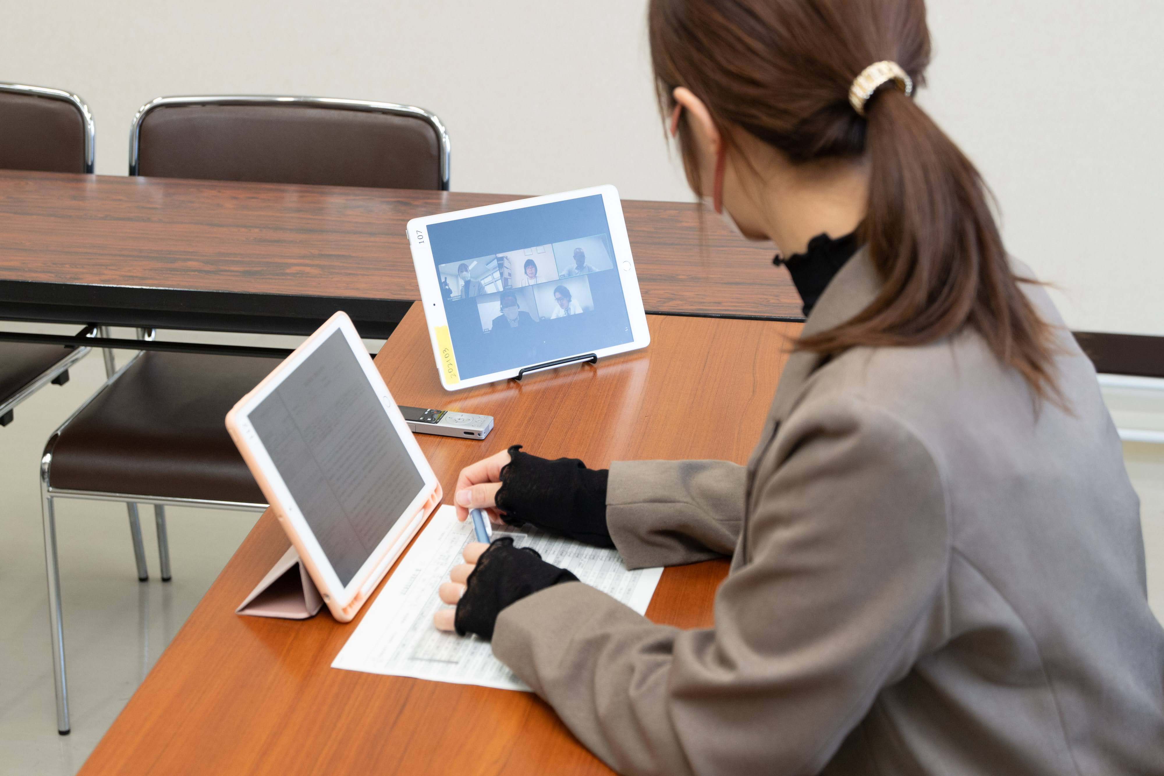 介護認定審査会をオンラインで開催する郡山市役所職員の様子。郡山市職員が会議室の机に着席し、審査会の委員が投影されているタブレットと、会議資料が投影されているタブレット2台を用いている。