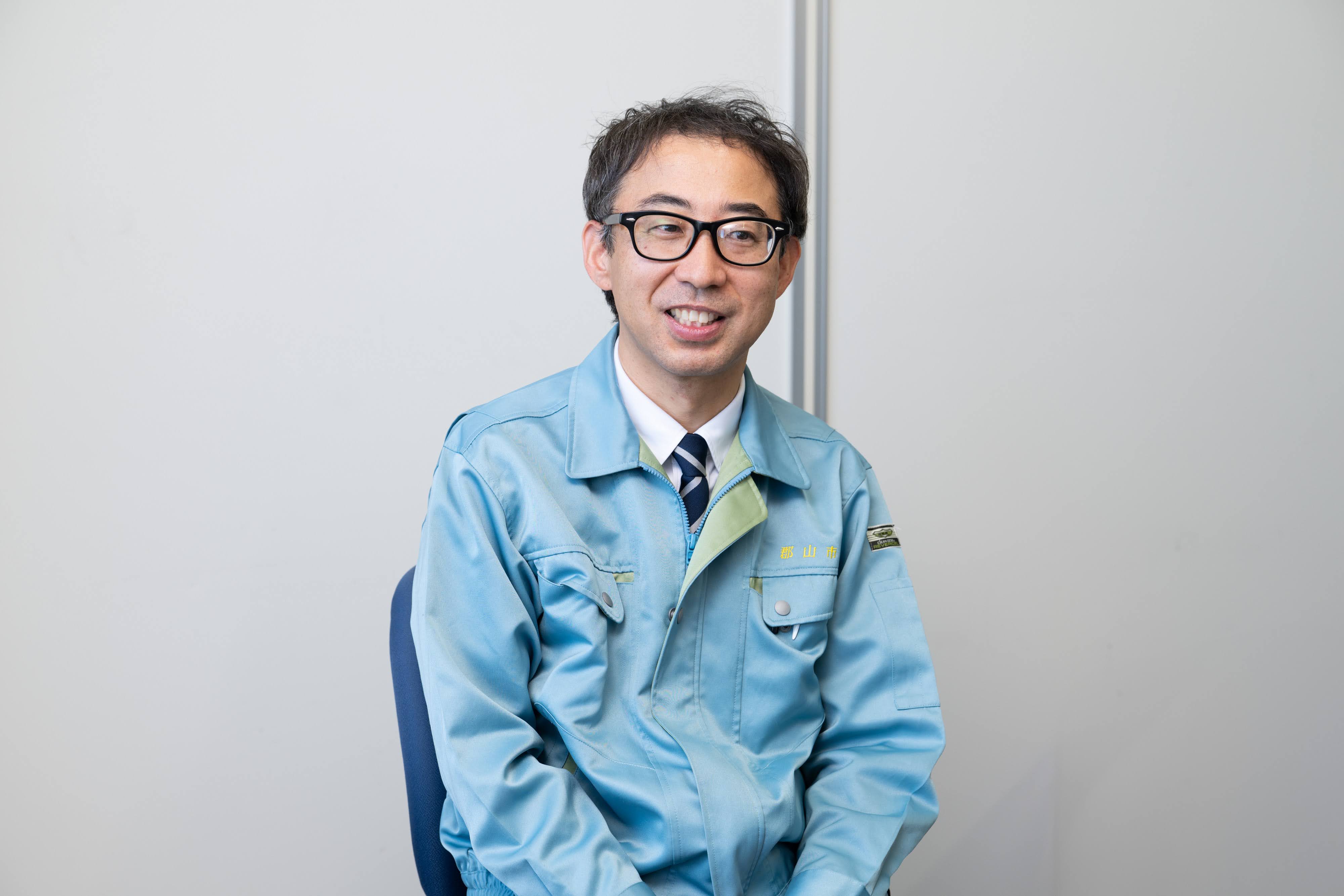 郡山市介護保険課の佐藤主任がインタビューに応じている写真。会議室の壁を背景に、椅子に腰かけている様子。