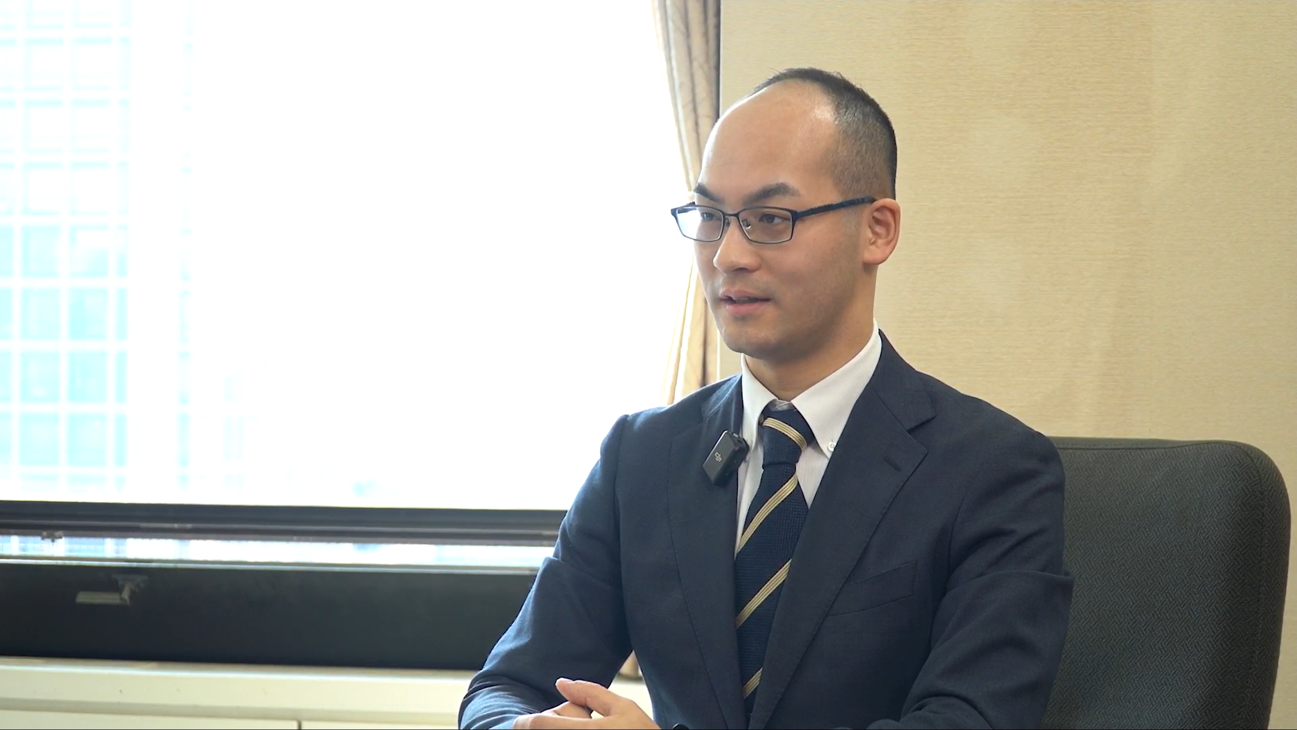 厚生労働省老健局老人保健課・渡井課長補佐がインタビューに答えている。会議室の壁と窓を背景に、着席している様子。