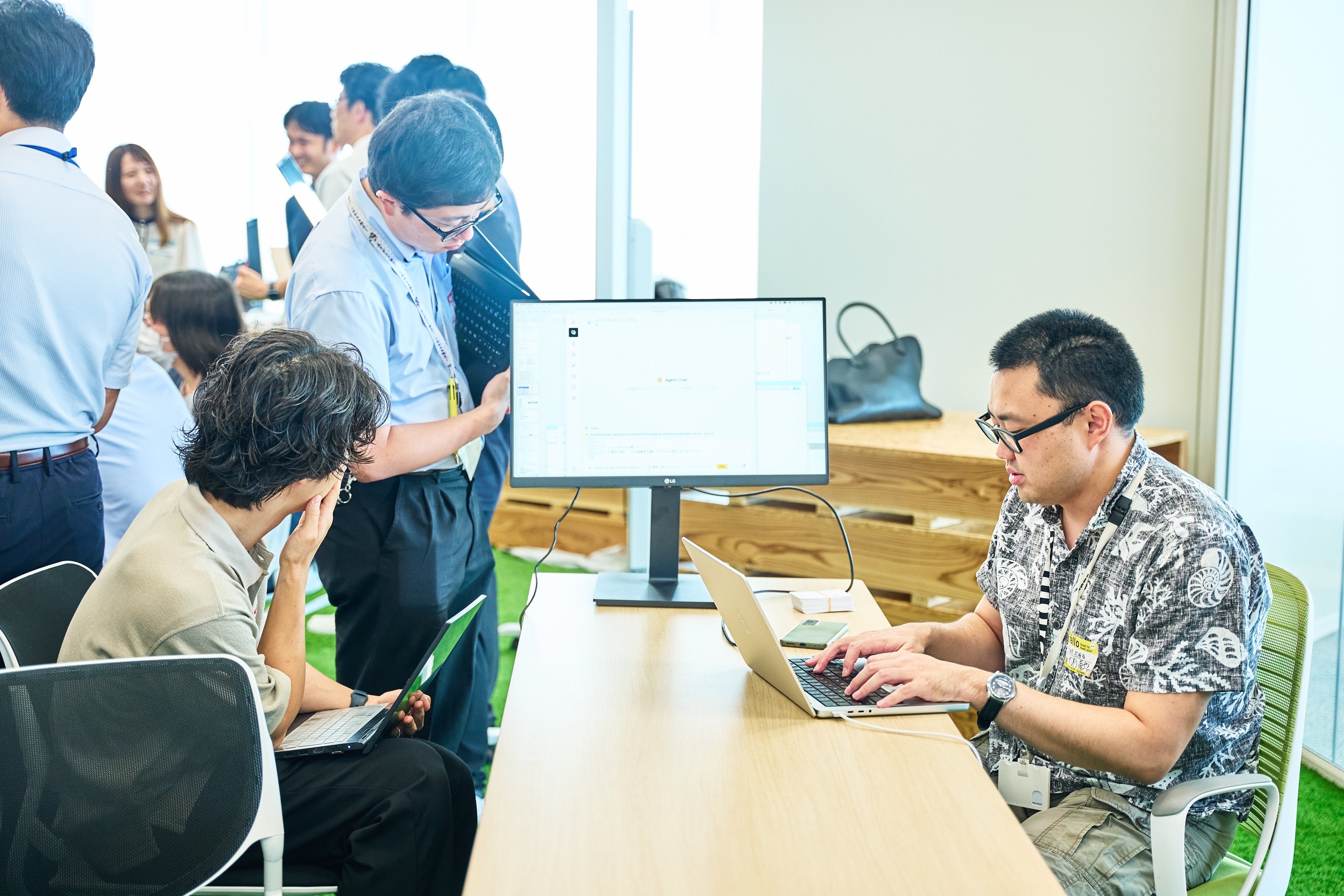 「共創PFキャンプinデジタル庁~自治体業務のAI活用編~」のワークショップの様子。着席しモニターにつないだパソコンを触っている人物、モニターを覗き込む2人の人物を横側から撮影している。