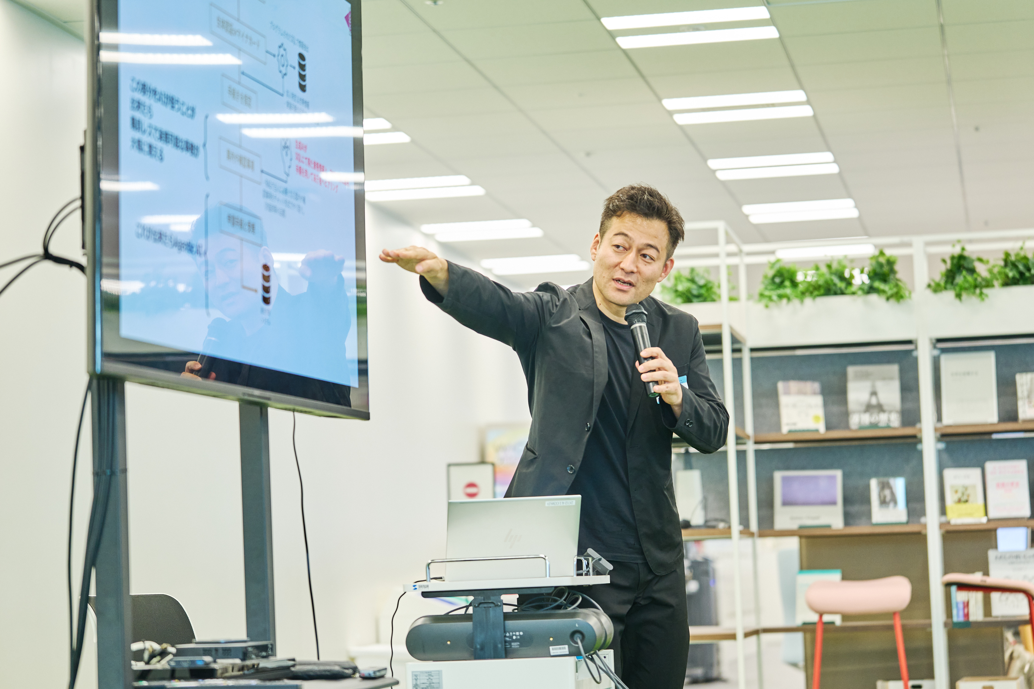 株式会社RKKCSの徳山泰之氏が登壇して発表している様子を正面から撮影。モニターを右手で指し示しながら、左手にマイクを持って話している。
