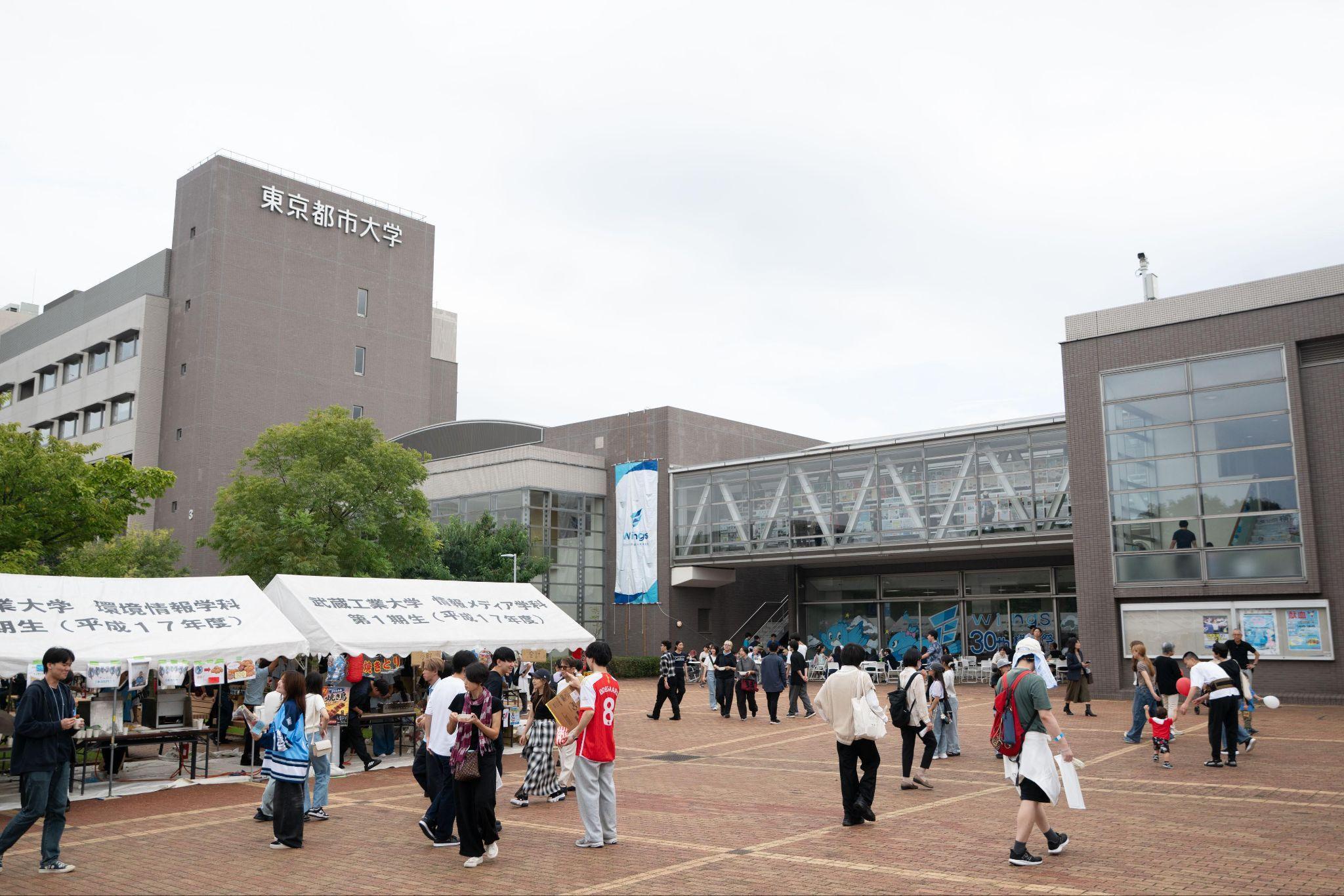 東京都市大学のキャンパスで開催された横浜祭の様子。広場には模擬店のテントが並び、多くの来場者が行き交っている。奥の建物には「Wings」の垂れ幕が掲げられている。