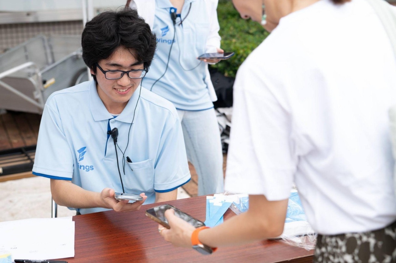 水色のポロシャツを着た学園祭スタッフの男性が、受付テーブルでスマートフォンを持ち、来場者のスマートフォンに表示された情報を確認している様子。