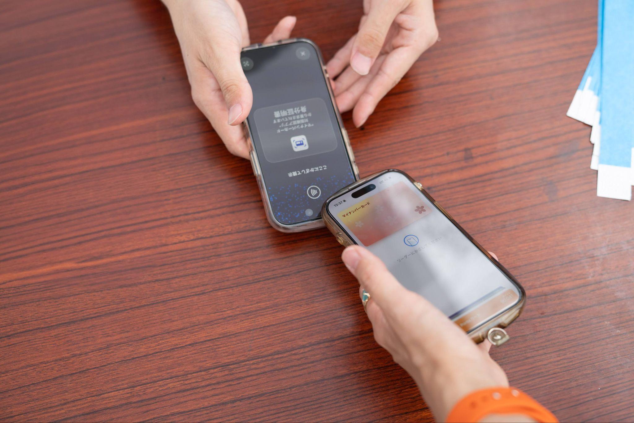 受付テーブルで、来場者がiPhoneのマイナンバーカードを、スタッフが持つ「身分証明書」読み取り画面のスマートフォンにかざしている様子。