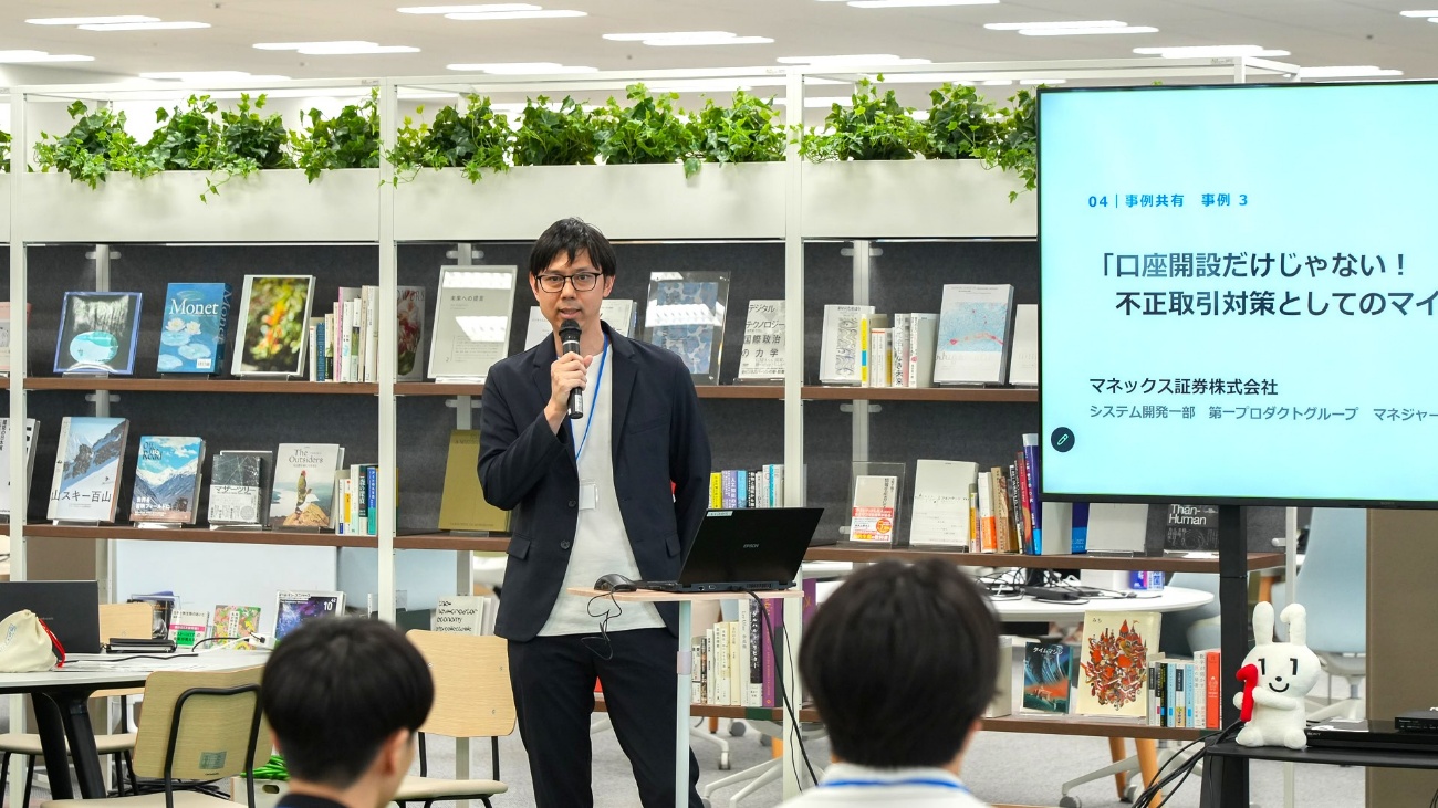 マネックス証券株式会社の法貴大輔氏がマイクを持って登壇している様子。法貴氏の右側にはスクリーンがあり、当日の資料の一部が投影されている。
