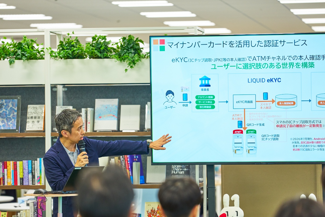 株式会社セブン銀行の三浦龍矢氏がマイクを持って登壇している様子。三浦氏の右側にはスクリーンがあり、「マイナンバーカードを活用した認証サービス」と題したスライドが映し出されている。話を聞いている参加者の後頭部が3人ほど確認できる。