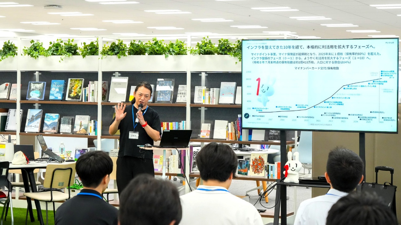 デジタル庁の小田部巧参事官補佐がマイクを持って登壇している様子。小田部参事官補佐の右側にはスクリーンがあり、「インフラを整えてきた10年を経て、本格的に利活用を拡大するフェーズへ」という文字が見える。また、小田部参事官補佐の話を聞く参加者の後ろ姿が5人ほど確認できる。