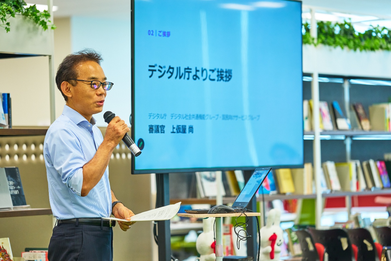 デジタル庁の上仮屋尚審議官がマイクと原稿を持って挨拶をしている様子。後ろのスクリーンには「デジタル庁よりご挨拶」「審議官 上仮屋尚」の文字が確認できる。