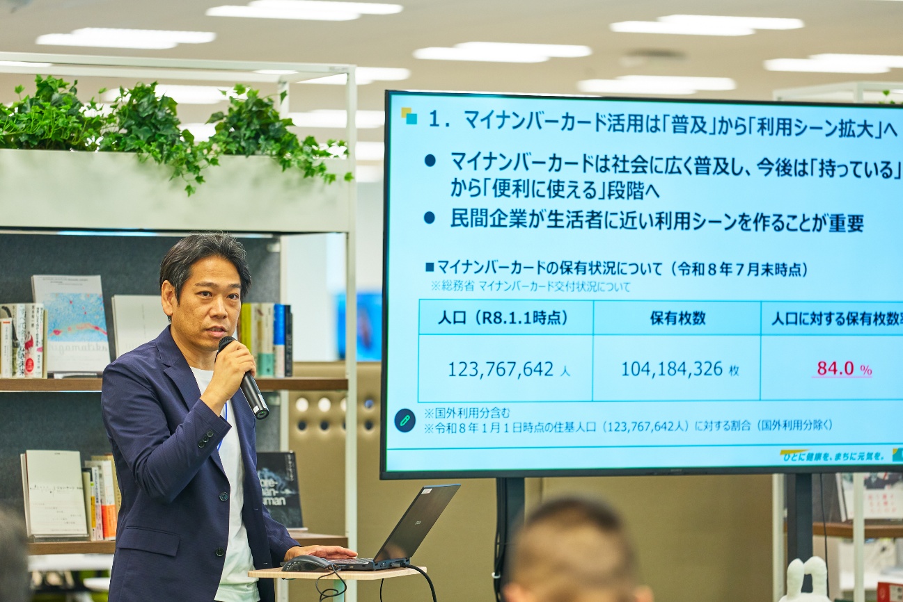 明治安田生命保険相互会社の小谷修一氏がマイクを持って登壇している様子を写した写真。小谷氏の右側にはスクリーンがあり、「マイナンバーカード活用は「普及」から「利用シーン拡大」へ」「マイナンバーカードは社会に広く普及し、今後は「持っている」から「便利に使える」段階へ」「民間企業が生活者に近い利用シーンを作ることが重要」といった文字が見える。また、マイナンバーカードの保有状況について（令和8年7月末時点）は、人口に対する保有枚数84.0%とも書かれている（人口123,767,642人、保有枚数104,184,326枚）。