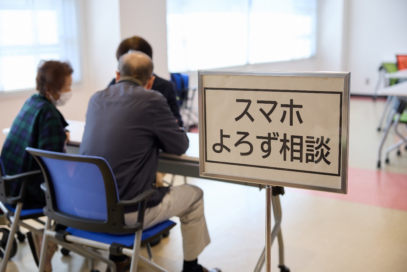 スマホよろず相談所の様子。 手前の右側に「スマホよろず相談」と大きく書かれたパネルスタンドの看板が設置されている。濃紺のジャケットに青・黒・白の幾何学模様のブラウス、メガネを着用した加賀市シニアスマホアンバサダー、デジタル推進委員の岸本博子さんが高齢女性と高齢男性がテーブル越しに向き合って着席している。