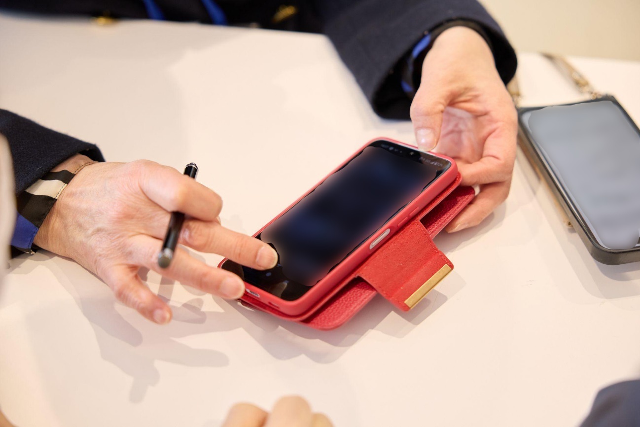 スマホよろず相談所の様子。 テーブルの上で、スタッフが右手にスタイラスペン（タッチペン）を持ち、横向きに置いたスマートフォンの画面に触れようとしている。スマートフォンは赤い手帳型スマートフォンケースに入っている。テーブル右端にはもう1台スマートフォンが置かれている。