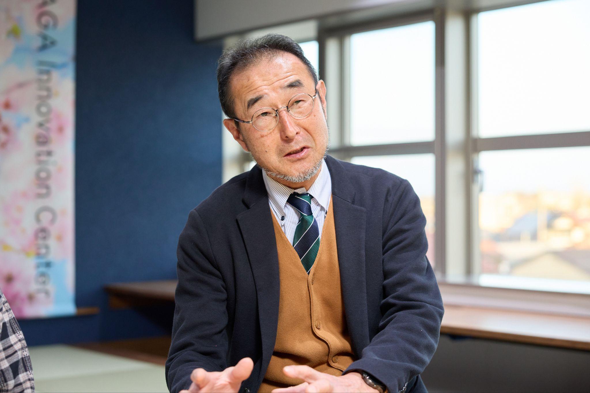 （加賀市シニアスマホアンバサダー、デジタル推進委員の立見征二さん。丸い眼鏡、濃紺のジャケット、キャメルのカーディガン、緑と紺のストライプネクタイを着用し、窓を背にしてインタビューに応じている様子。）