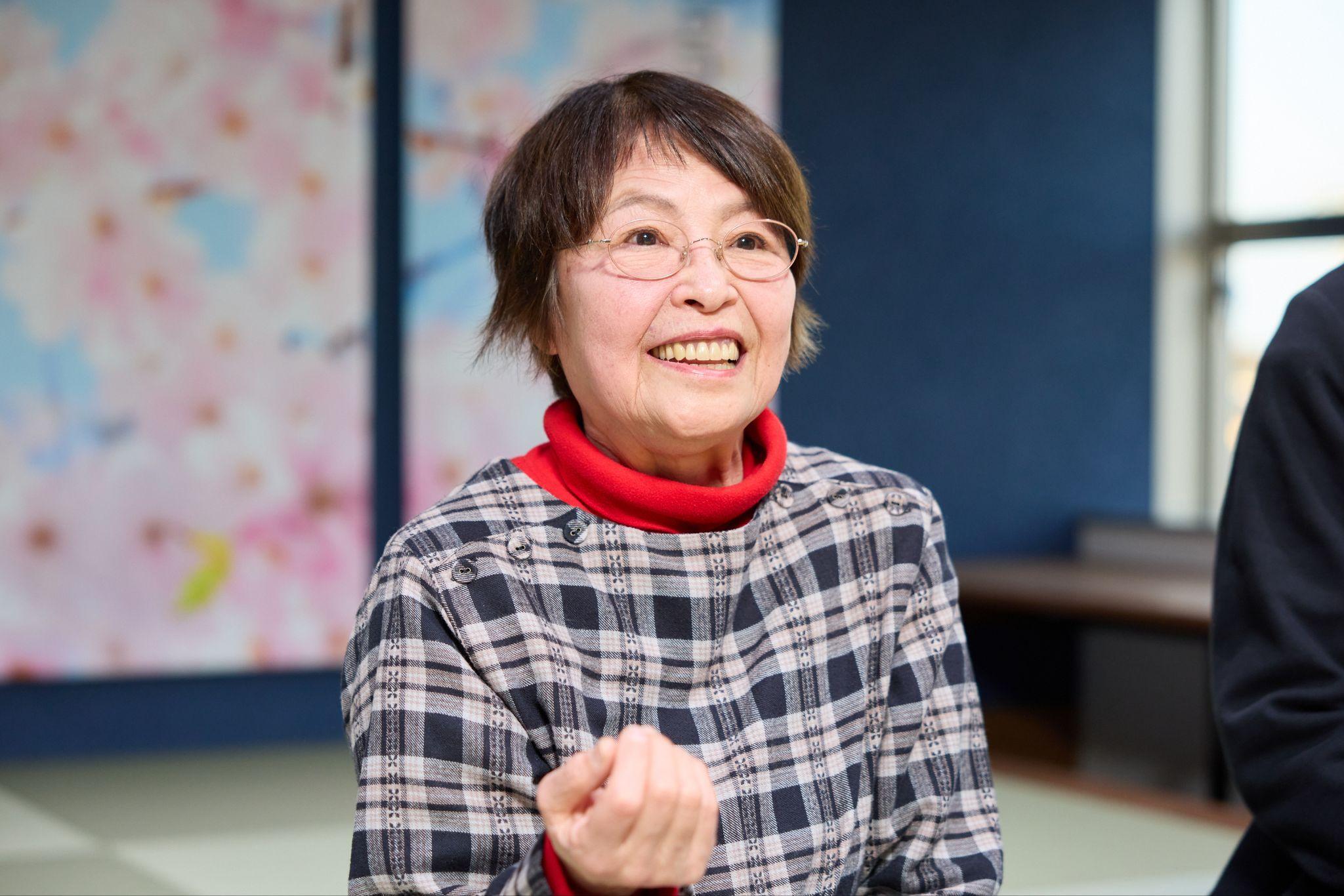 （加賀市シニアスマホアンバサダー、デジタル推進委員の山野ちさ子さん。丸い眼鏡、赤いタートルネック、グレーのチェック柄トップスを着用し、窓を背にしてインタビューに応じている様子。）