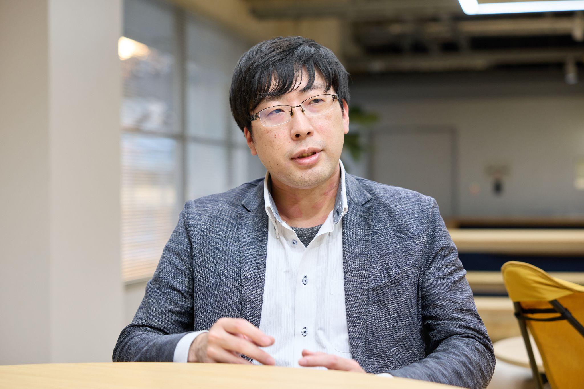 スマホよろず相談所で講師を務める株式会社インテトラスの村中氏。ハーフリムの眼鏡、グレーのジャケットに白いシャツを着用し、テーブルに座ってインタビューに応じている。背景には窓、机がある。