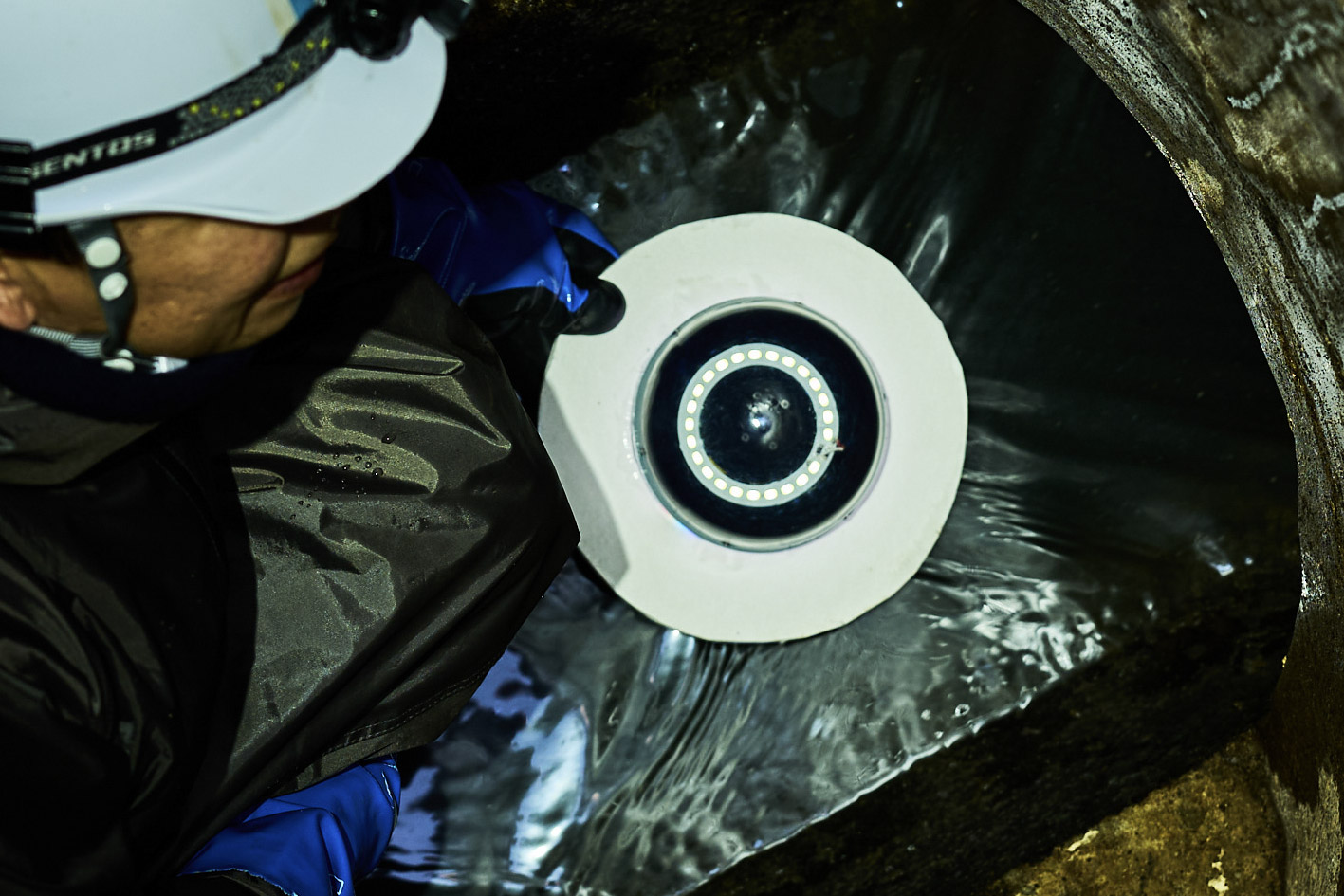 ヘルメットを着用した作業員が下水道の水流に球体型カメラ装置を載せようとしている様子を真上から撮影している。