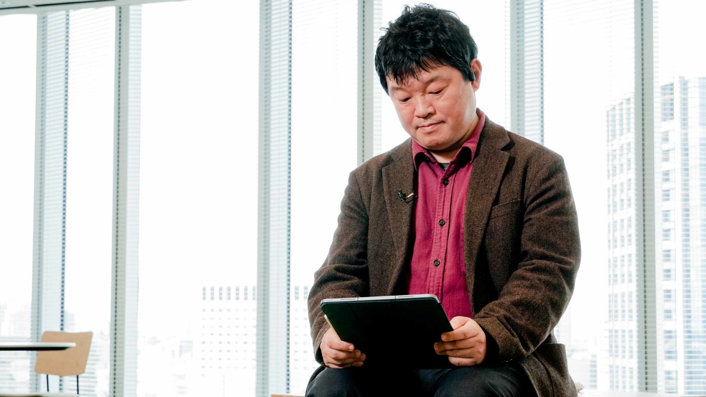 窓越しの椅子に座りタブレットを見ている楠、エンジのシャツに茶のジャケットを着ている姿。