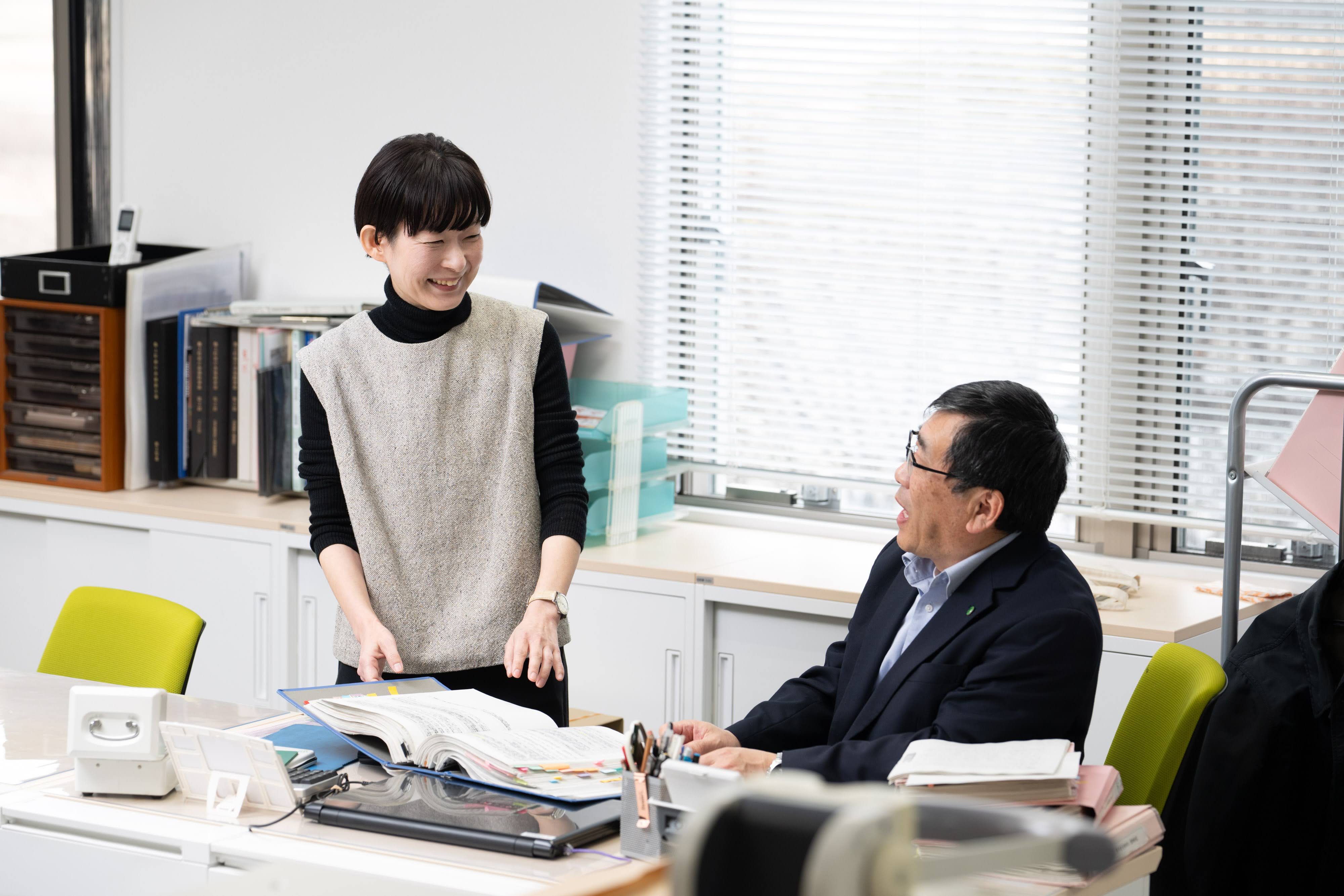 築上町企画財政課の舛川係長（左）と椎野課長が談笑している様子。椎野課長の自席にて、椎野課長が着席し、舛川係長がその傍らで資料が綴じられたファイルを見せている。