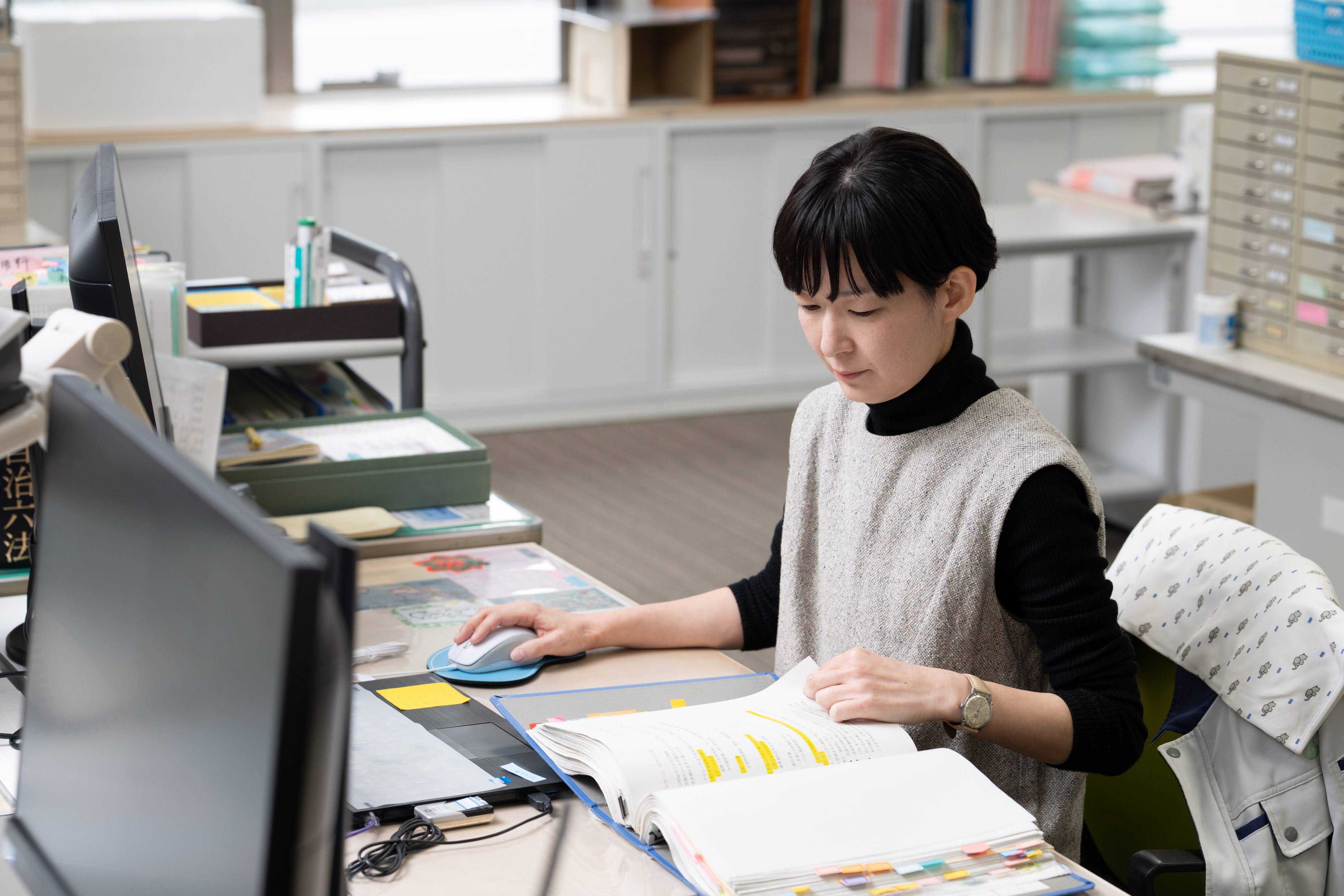 築上町企画財政課の舛川係長が執務スペースの自席に着席し、紙の資料を左手でめくりながら、右手にマウスを操作している様子。