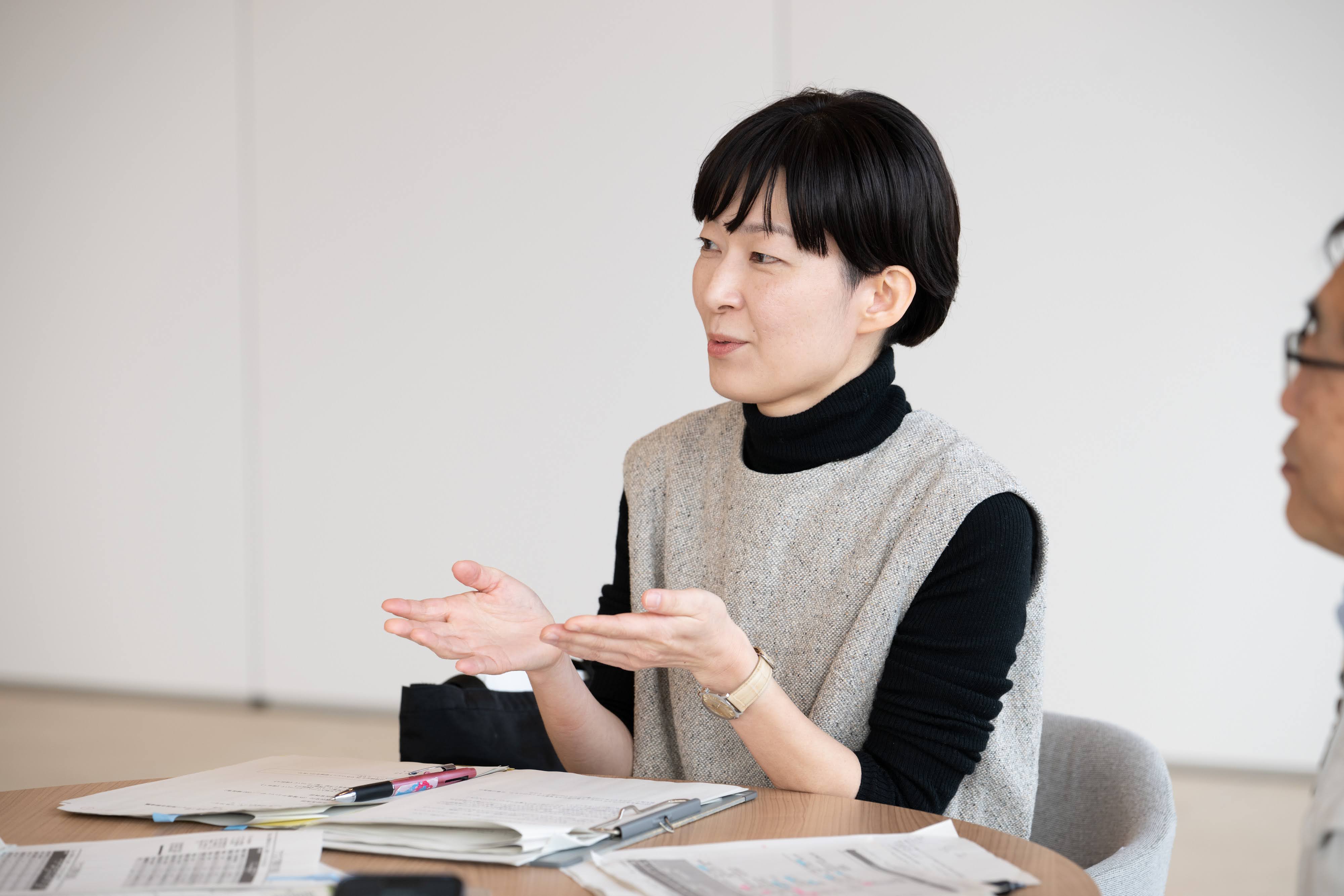 築上町企画財政課の舛川係長がインタビューに答えている様子。着席し手振りを交えながら話す様子を、正面向かって右手から撮影している。椎野課長が右側に写り込んでいる。