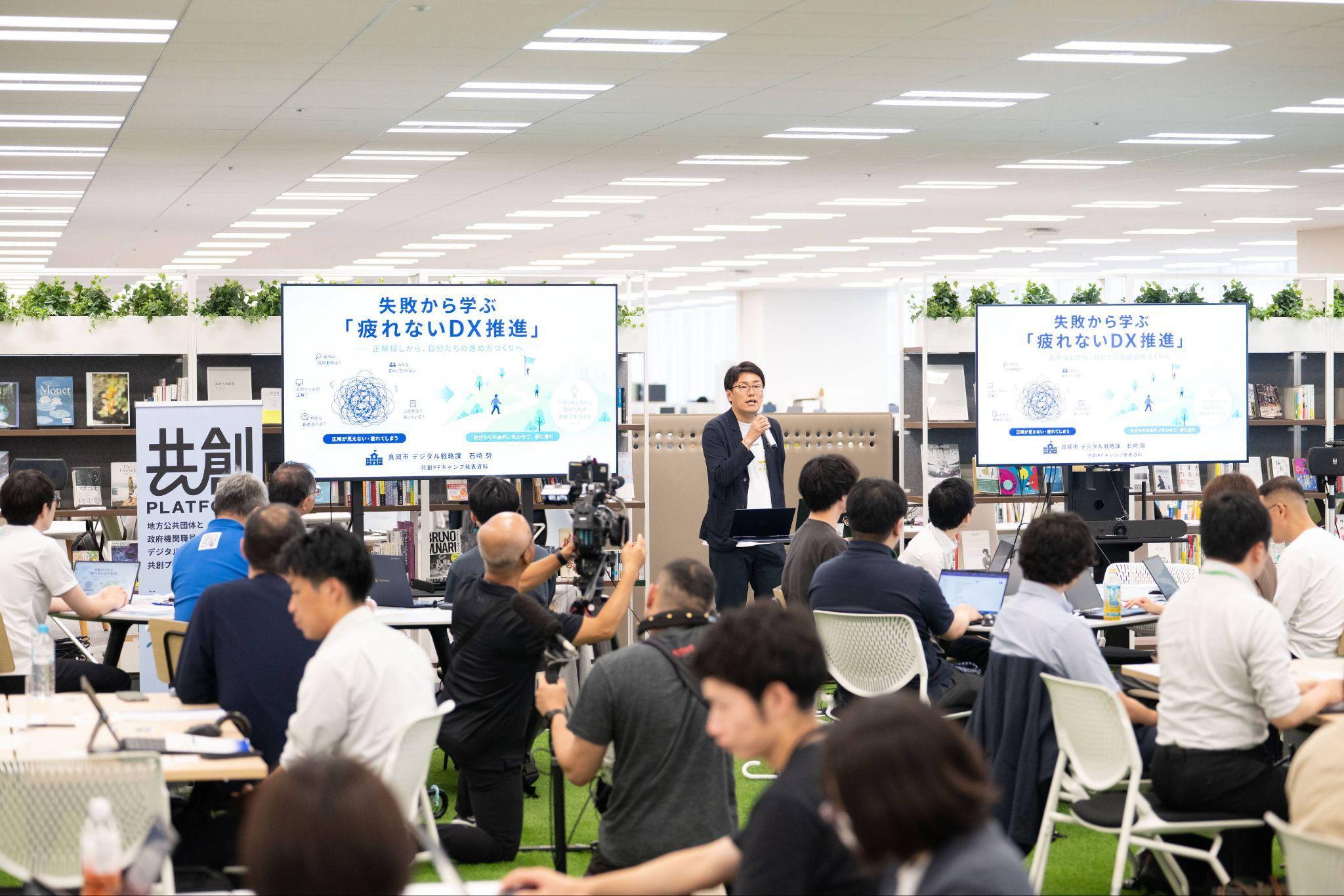 栃木県真岡市の担当者である石崎努氏が右手にマイクを持って、登壇している様子の写真。石崎氏の隣にはモニターが設置されており、「失敗から学ぶ「疲れないDX推進」」と書かれているスライドが映されている。参加者が石崎氏の話を聴きながら、モニターを見ている後ろ姿も写されている。イベント自体はデジタル庁で行われたが、デジタル庁には芝生エリアと呼ばれる広いスペースがあり、いろいろなテーマに合わせた本が置かれている。その本の一部も写真に背景として写っている。