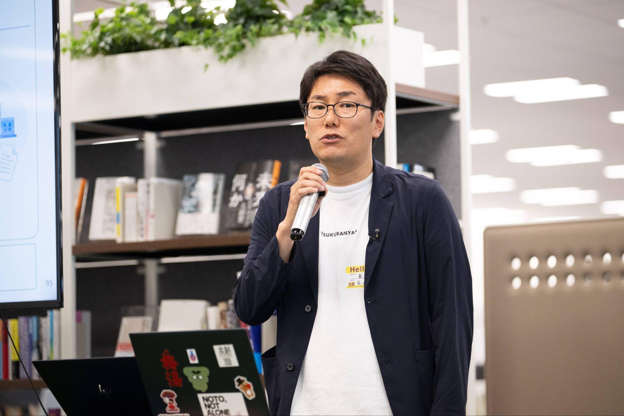 登壇する栃木県真岡市役所の石崎努氏。紺色のジャケットに、白いシャツを着て、右手にマイクを持っている。目の前にはノートパソコンが2台置いてある。イベント自体はデジタル庁で行われたが、デジタル庁には芝生エリアと呼ばれる広いスペースがあり、いろいろなテーマに合わせた本が置かれている。その本の一部も写真に背景として写っている。