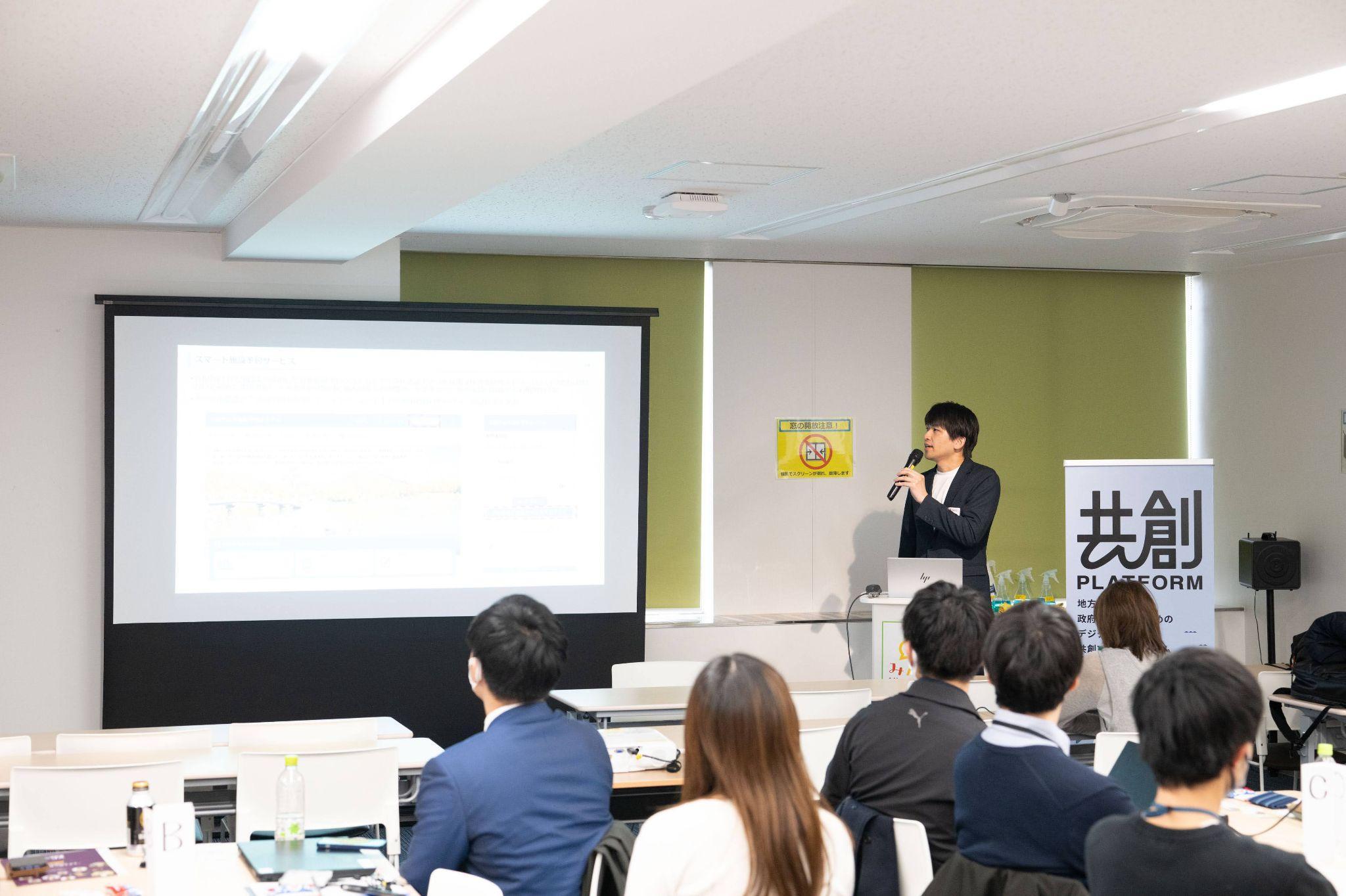 兵庫県三田市総務部DX推進課スマートシティ推進係の岩﨑謙二係長がマイクを持ってプレゼンテーションしている様子。