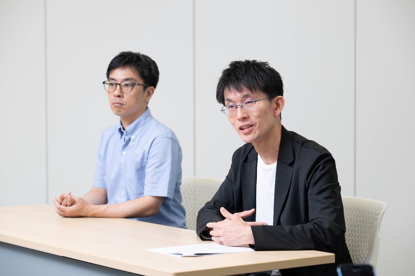 インタビューに応じるデジタル庁の黒籔参事官と経済産業省の木村補佐の写真。オフィスで壁を背景に着席し、机の上に両手を置いて話している様子を左斜め前から撮影している。