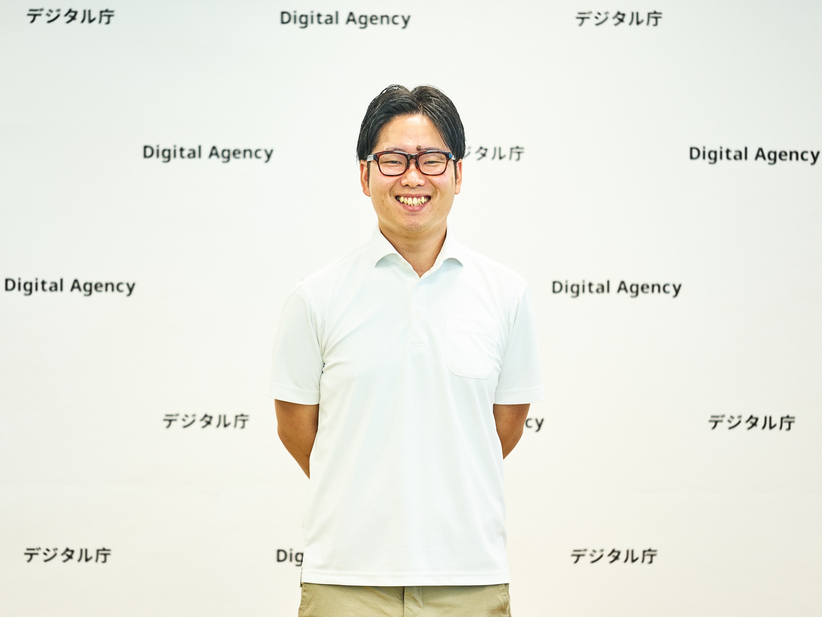 デジタル庁（Digital Agency）のロゴが繰り返し印刷された白いバックパネルを背景に、男性が正面を向いて立っている。両手を後ろに組み、笑っている写真。