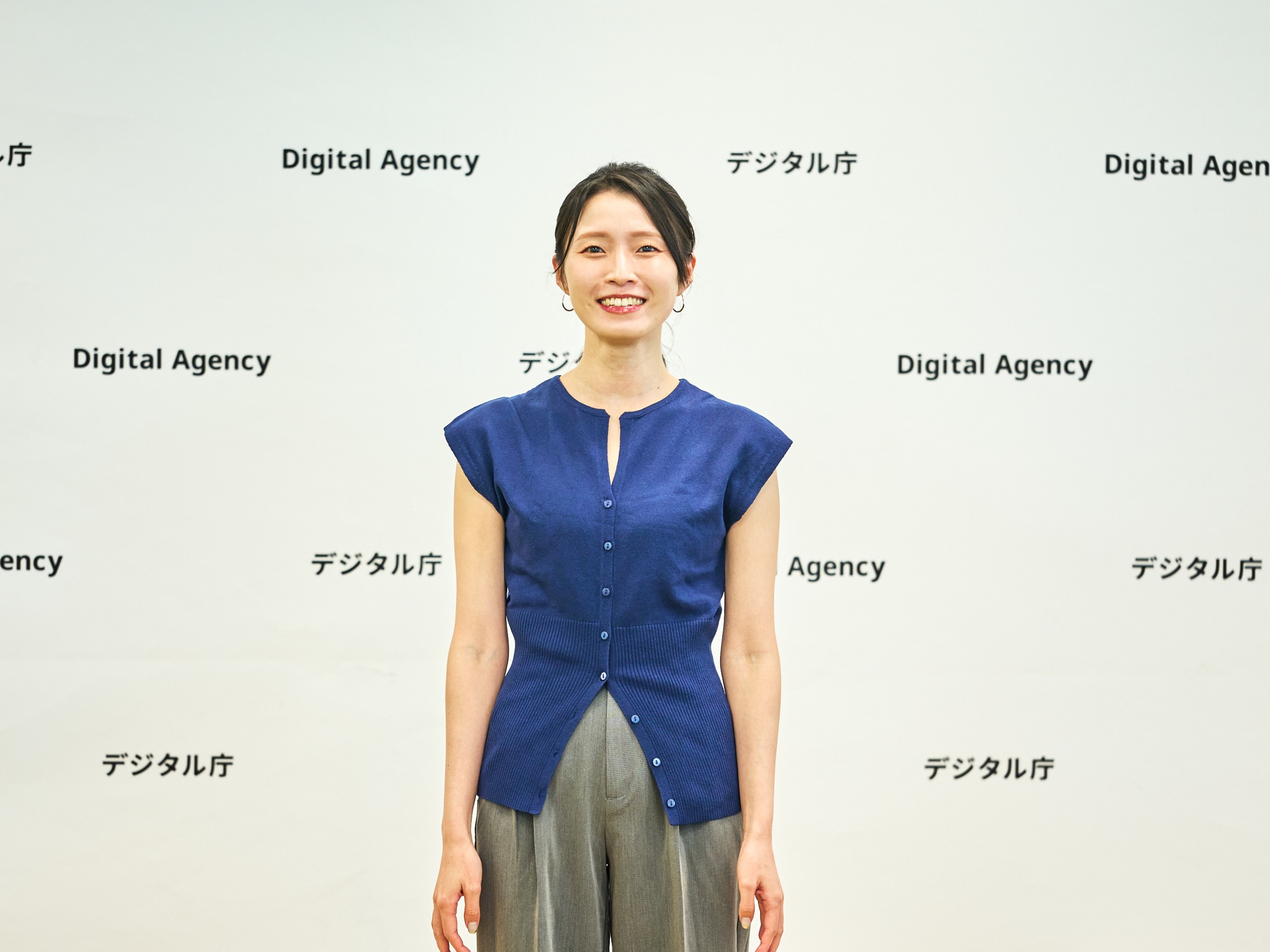 デジタル庁（Digital Agency）のロゴが繰り返し印刷された白いバックパネルを背景に、女性が正面を向いて立っている。両手を体の脇に自然におろし、笑顔を見せている。