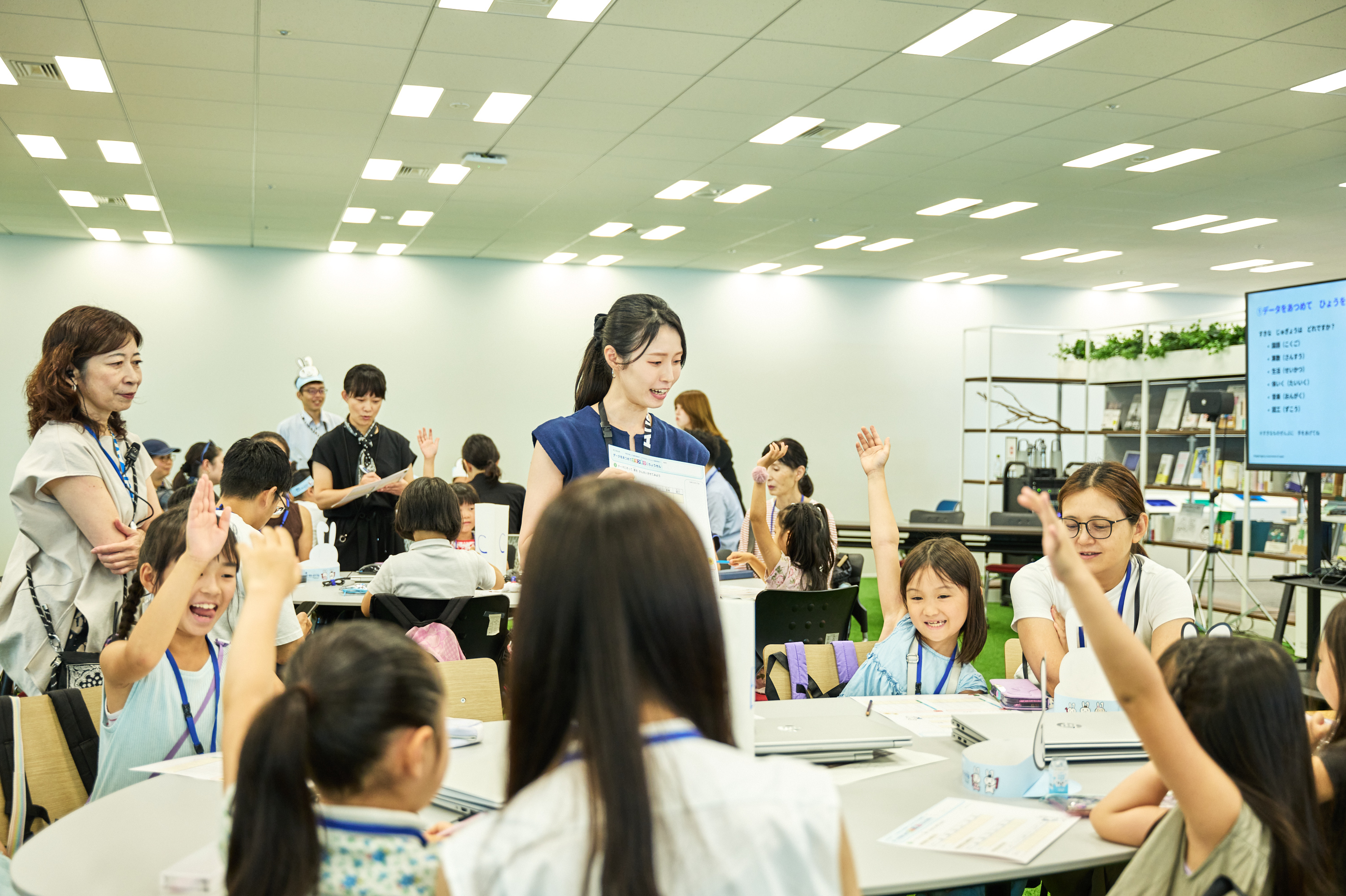 小学生の子どもたちが複数のテーブルに分かれてノートパソコンを前に、テーブルを囲むように座っている。手前のテーブル中央に立つ若い女性ファシリテーターが笑顔で話しかけており、多くの子どもが元気よく手を挙げている。右奥のモニターには『データをあつめて ひょうを』という文字が映し出されている。床には人工芝が敷かれ、本棚が置かれた空間で開催されている。