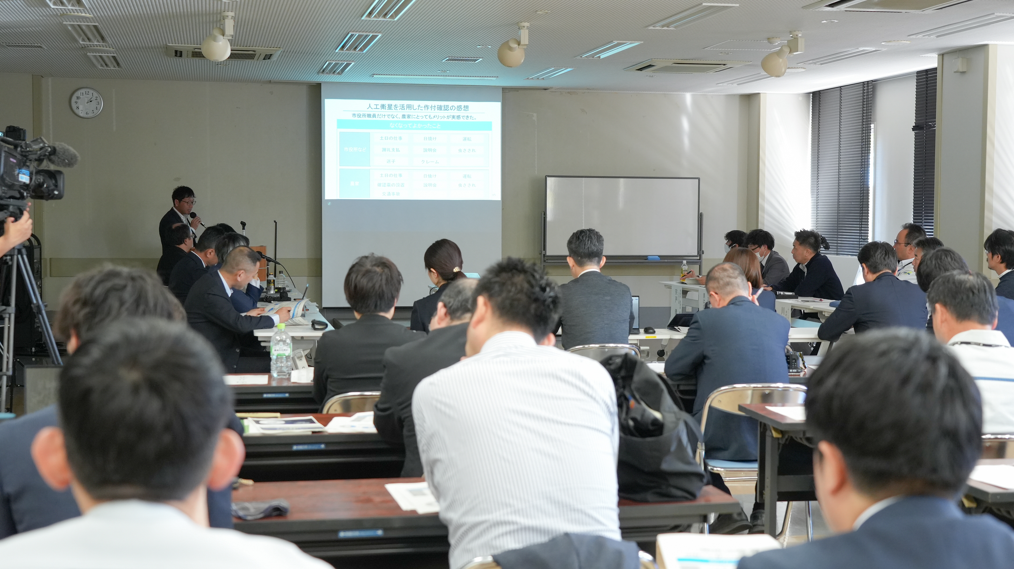 南相馬市が開催した成果報告会の会場の様子。会場後方から撮影。中央に資料が投影されたスクリーンがあり、スクリーンの左手に発表者が立って発言している。会場前方には他の発表者ら、会場後方には参加者が着席している。