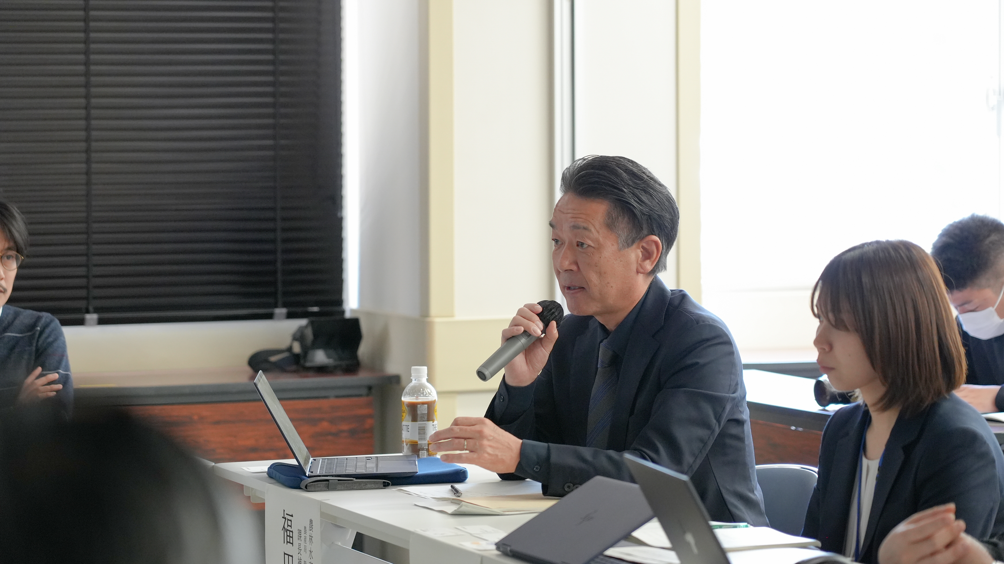 報告会で発言する福田課長補佐を、左手から撮影した画像。着席した状態で、マイクを持ち発言している。