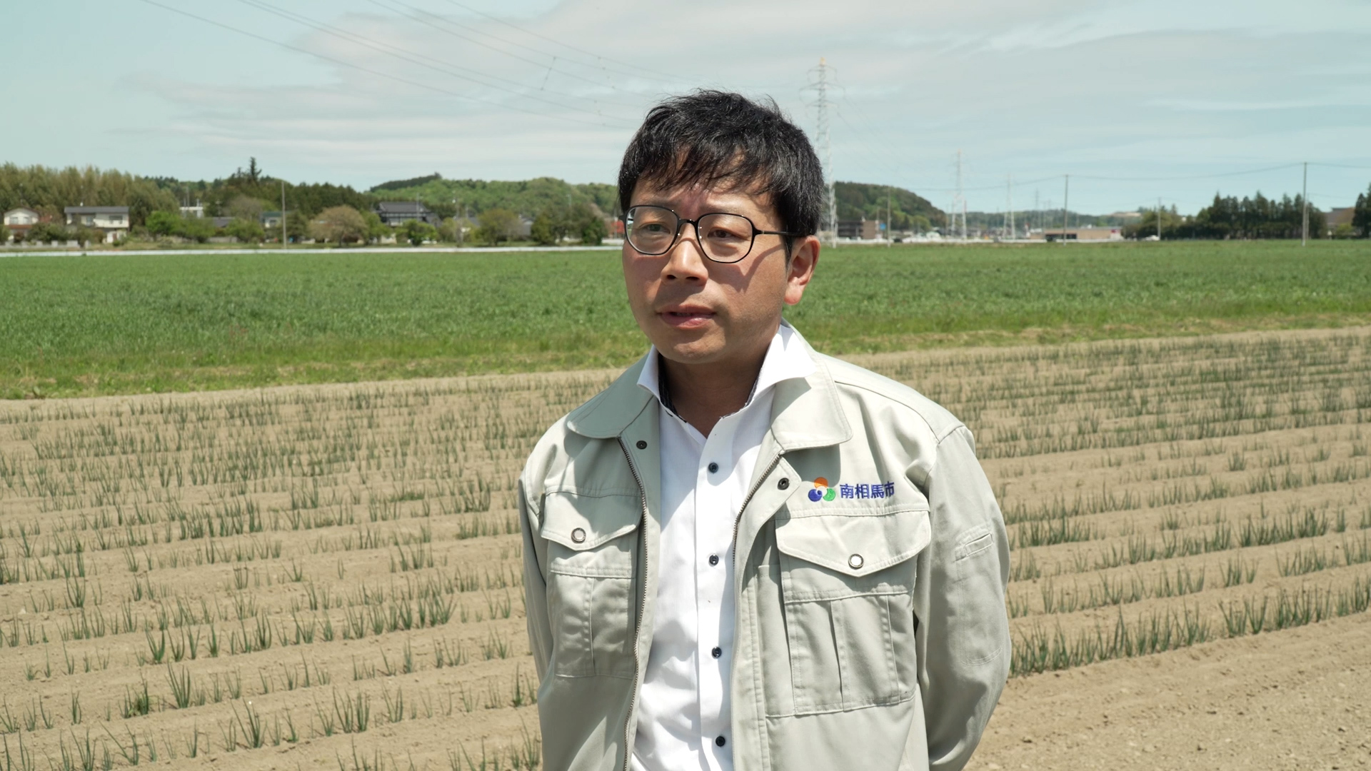 農地を背景に立つ、南相馬市農政課の大谷課長補佐のバストアップ写真。