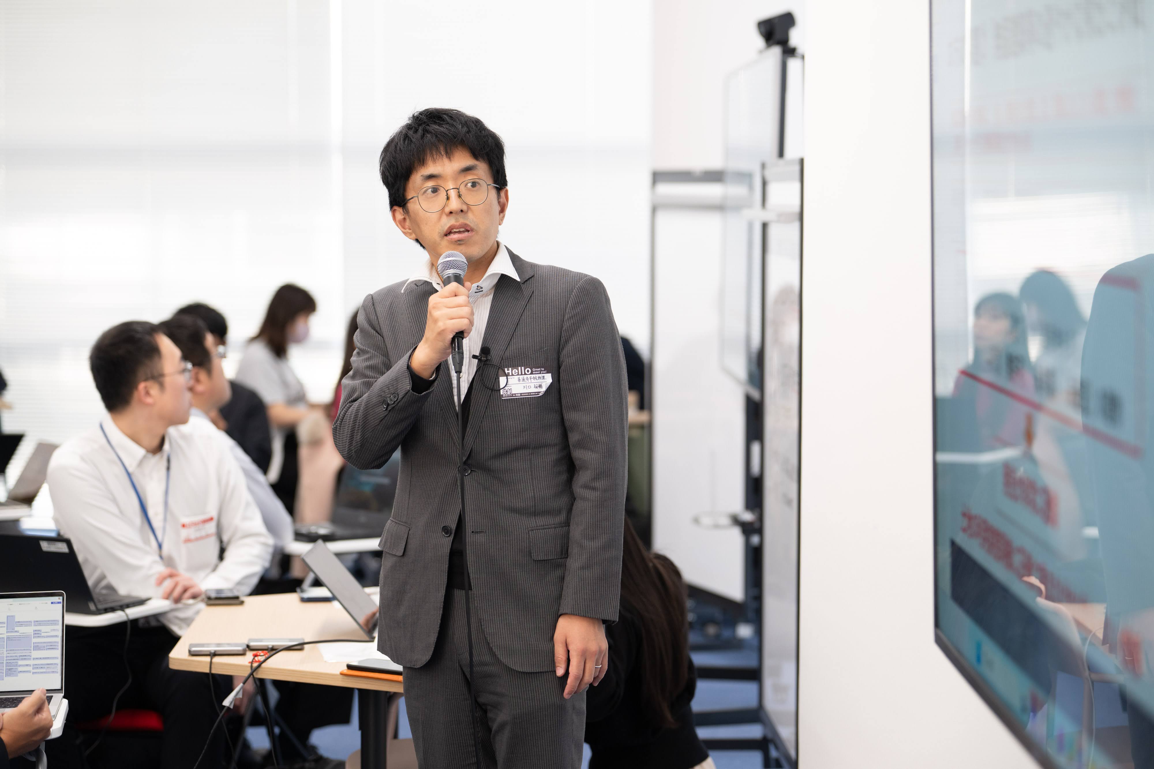 善通寺市の川口氏がマイクを持って、右側に映し出されたスクリーンを見ながらプレゼンテーションをしている様子。