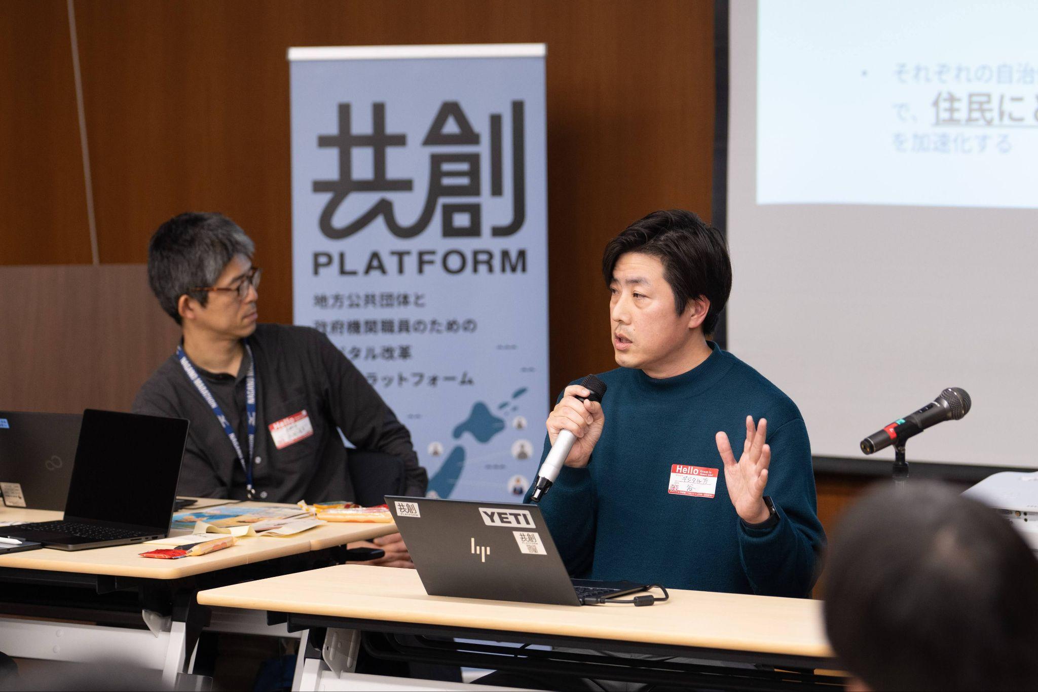 イベント会場でマイクを持ち発言するデジタル庁デジタル社会共通機能グループ窓口DX推進チームの谷吉郎参事官補佐。濃い緑色のセーターを着用し、マイクを持ちながら右手を挙げて話している。机の上にはノートパソコンが置かれている。隣には黒いジャケットを着用し、眼鏡をかけた男性が同席している。背後には『共創 PLATFORM 地方公共団体と政府機関職員のためのデジタル改革プラットフォーム』と書かれたロールアップバナーが立てられており、右奥にはプロジェクタースクリーンが映し出されている。