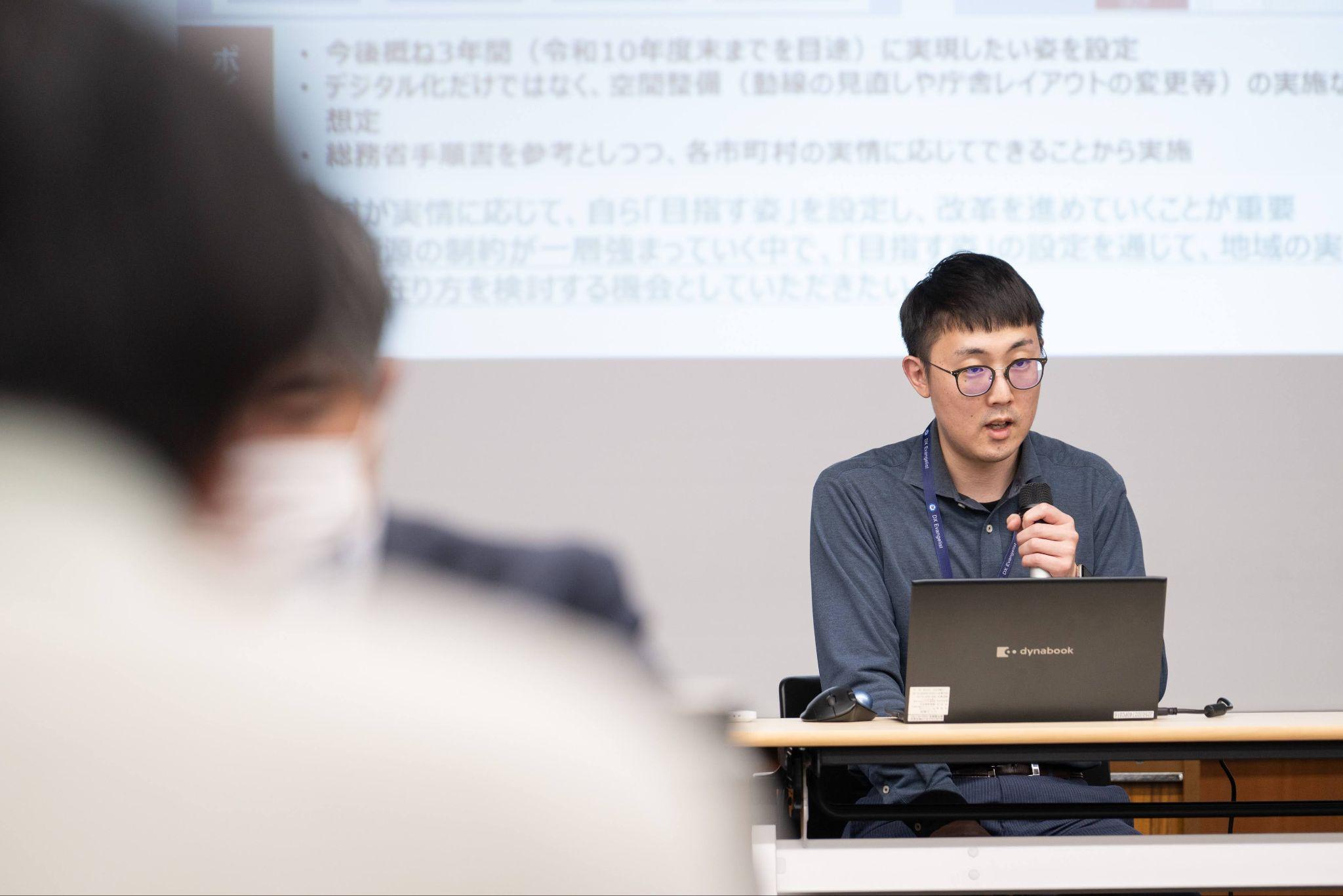 イベント会場でノートパソコンの前に座り、マイクを持って説明する長野県企画振興部DX推進課スマート自治体推進係の川口友輔氏。グレーの長袖シャツを着用し、机の上にはノートパソコンとマウスが置かれている。背後のスクリーンにはフロントヤード改革やデジタル化に関する資料が投影されている。