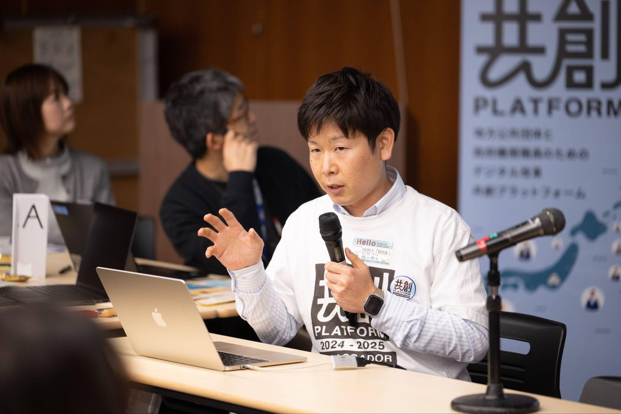 静岡県裾野市健康福祉部総合福祉課地域福祉係の勝又優斗氏が右手にマイクを持ちながら発言している写真。共創PLATFORM 2024-2025 AMBASSADOR」と書かれたTシャツを着用している。手元にはAppleのMacBookが開かれており、机の上には資料が置かれている。背後には「共創 PLATFORM」と書かれた縦型バナーが見え、右奥にはマイクスタンドが確認できる。