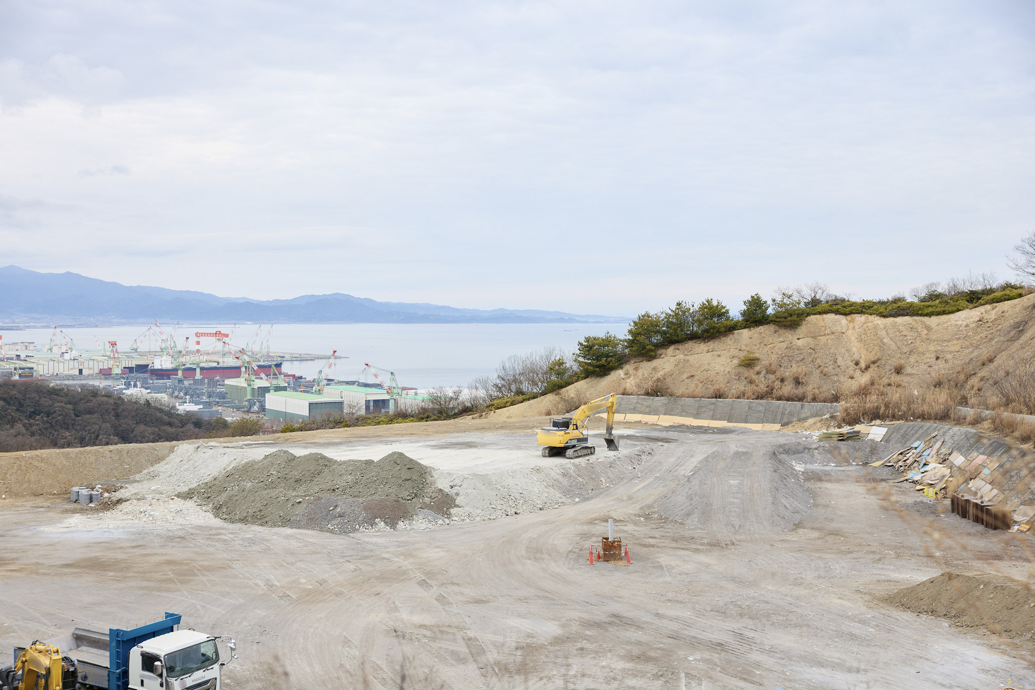 検査対象となった産業廃棄物最終処分場を撮影した写真。手前には砂利や土砂の山が積まれ、黄色いショベルカーが中央付近にある。左下にはトラックが見える。背景にはクレーンが立ち並ぶ港湾エリアと、遠くに山並みが広がる。空は薄曇りで明るい。