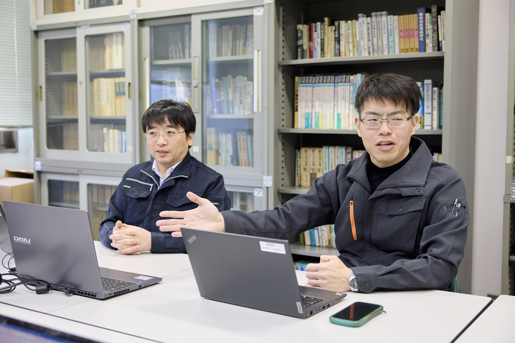 橋本廃棄物指導係長（左）と角田担当係長が白いテーブルに並んで座っている。角田氏が手ぶりを交えて話している。テーブルにはそれぞれノートパソコンが置かれており、スマートフォンも置かれている。背後には書棚が並んでいる。