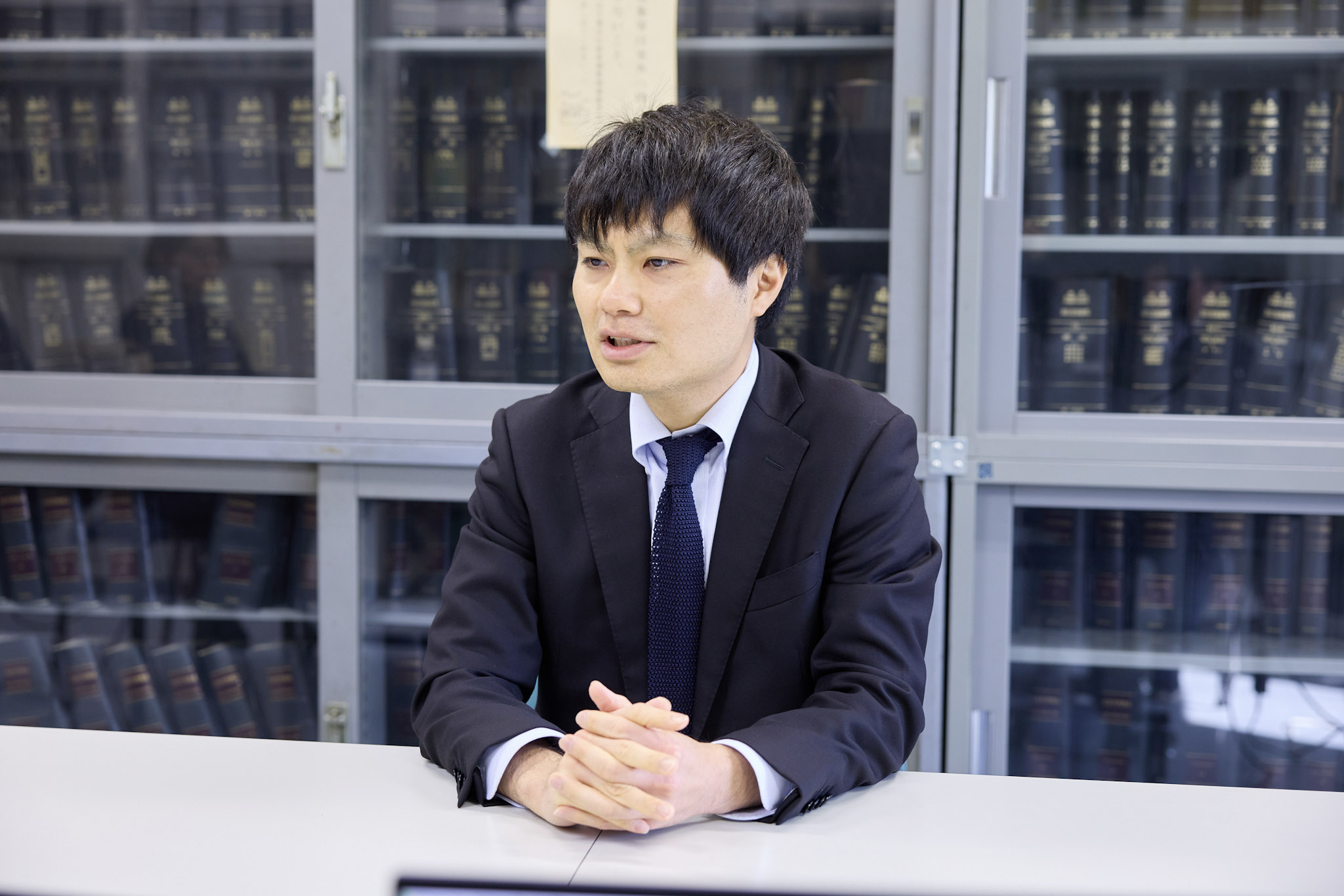 環境省の浅利達郎・参事官補佐が白いテーブルの前に座り、両手の指を組んでやや前傾みの姿勢で話している様子を正面斜め右前から撮影している。背景には濃紺の背表紙の書籍が並んだガラス扉付きの書棚が広がる。