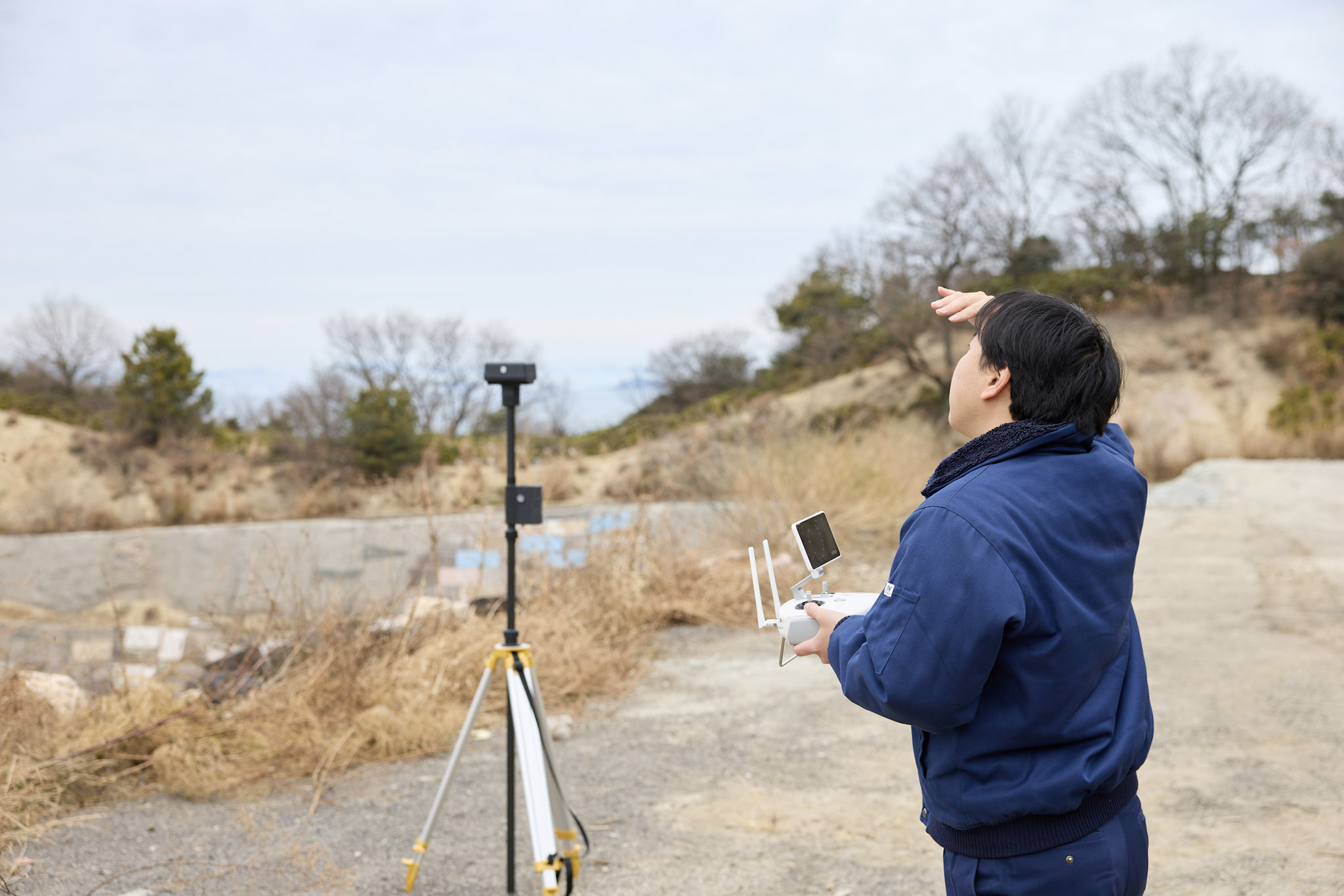 屋外にて、紺色の作業着を着た男性が左手でドローンのプロポ（送信機）を持ちながら、右手を額に当てて上空を見上げている。男性の左側にはセンサーをマウントした三脚が設置されている。