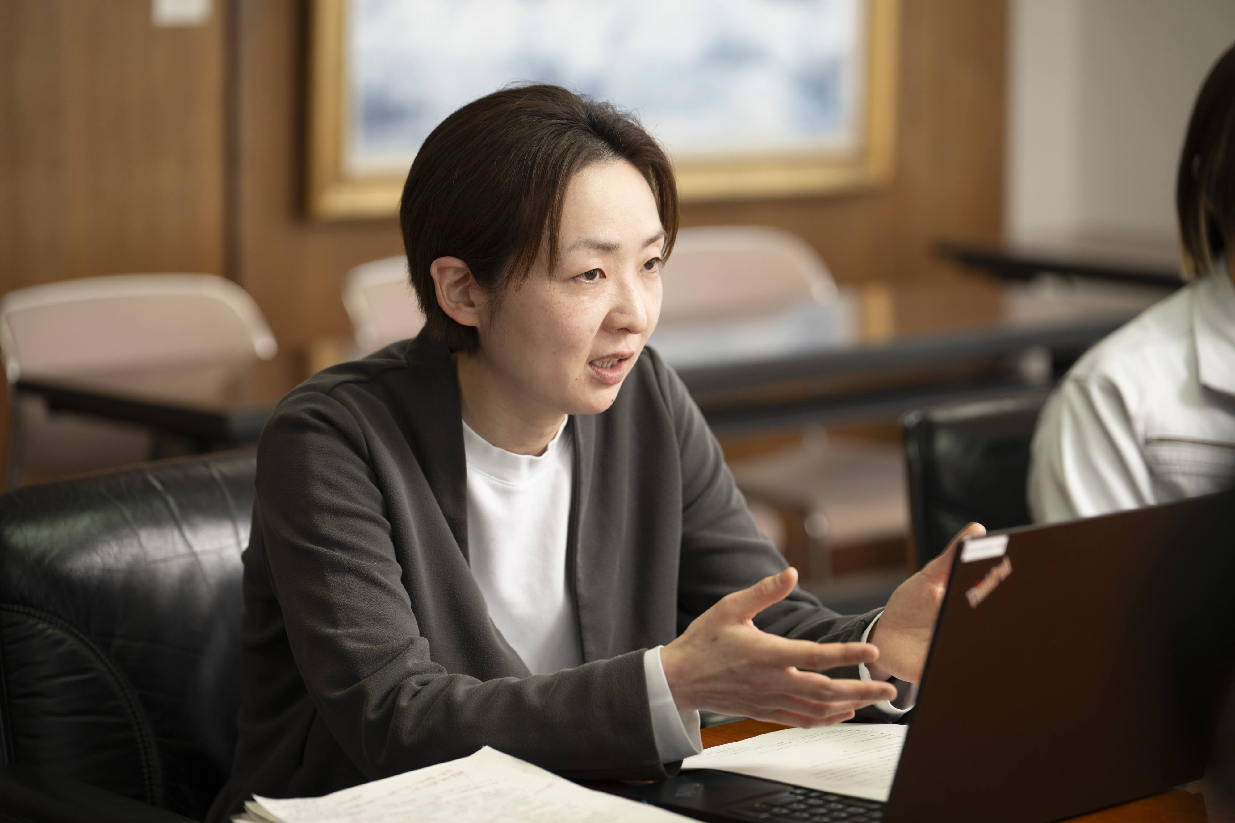 会議室でノートパソコンの前に座り、身振りを交えながら話す田川市経営企画課企画政策係の陸田主任。木目調の壁と絵画が飾られた会議室で、黒いノートパソコンの前に座っている。右手を前に出して身振りを交えながら、やや右方向を向いて話している。