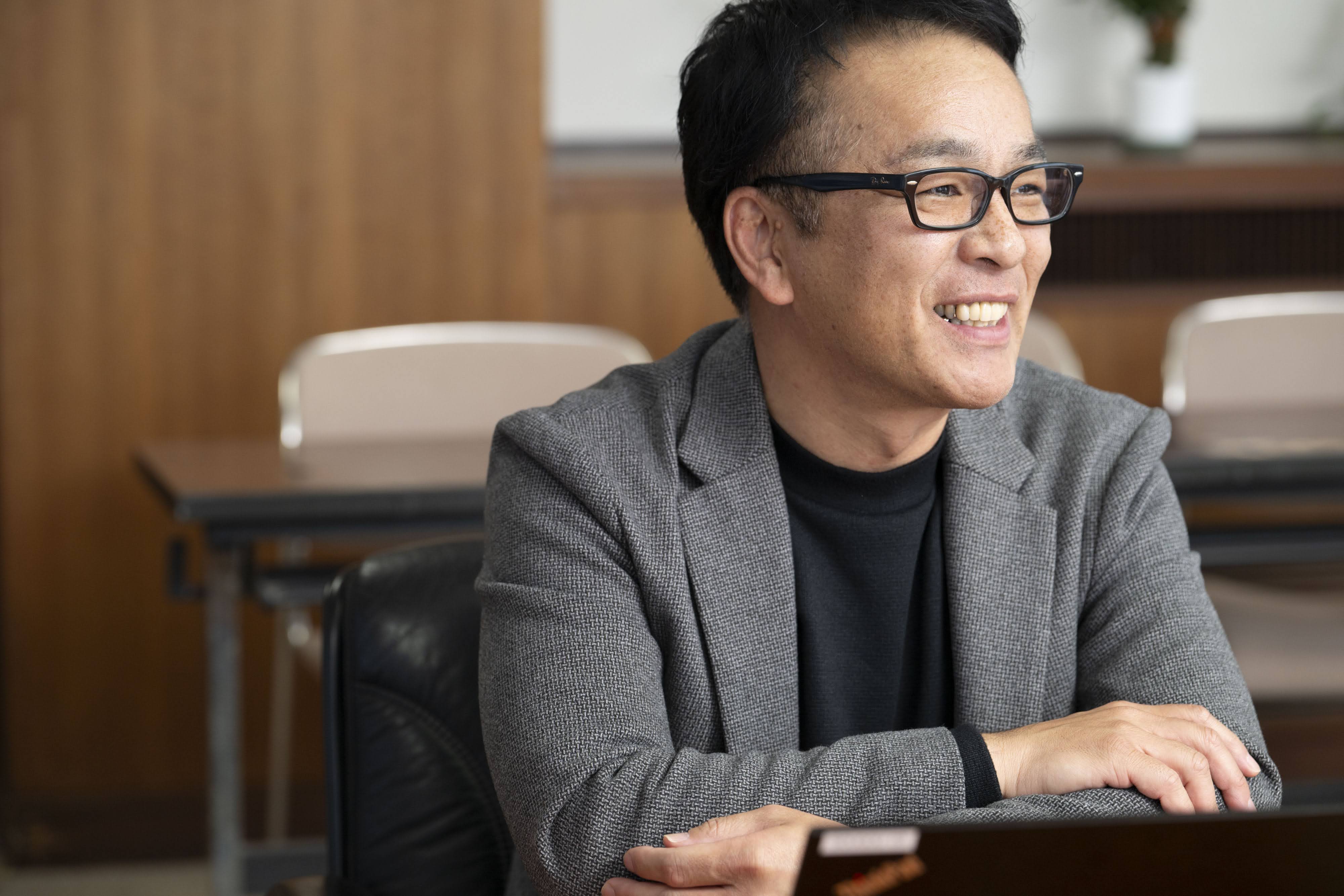 会議室で腕を組み話す、経営企画課の有田係長。木目調の壁が背景の会議室で、視線はやや左方向に向けられている。