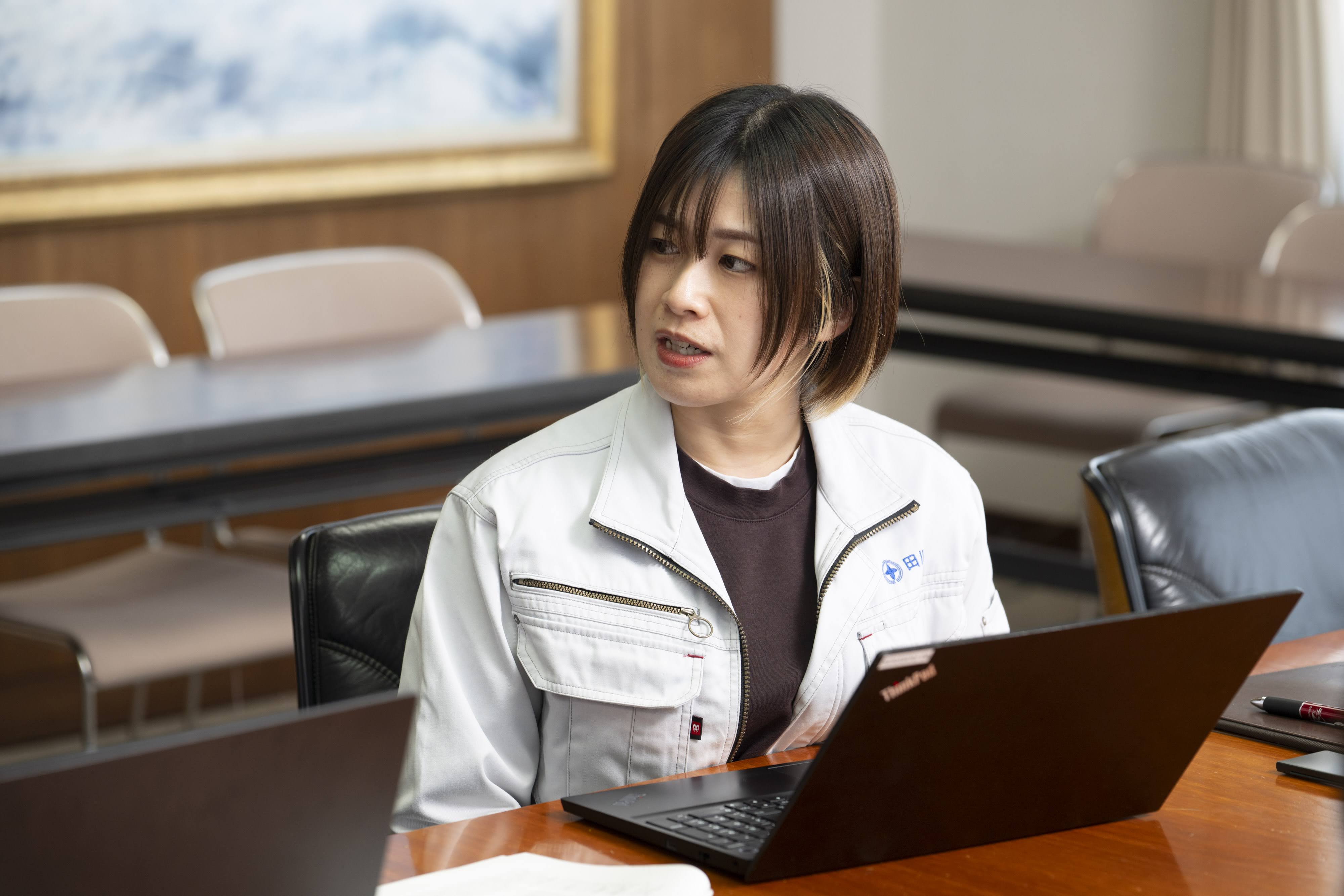 会議室でノートパソコンの前に座り、身振りを交えながら話す、田川市経営企画課DX推進室の三根氏。 木目調の壁と絵画が飾られた会議室で、黒いノートパソコンの前に座っている。右手を前に出して身振りを交えながら、やや右方向を向いて話している。