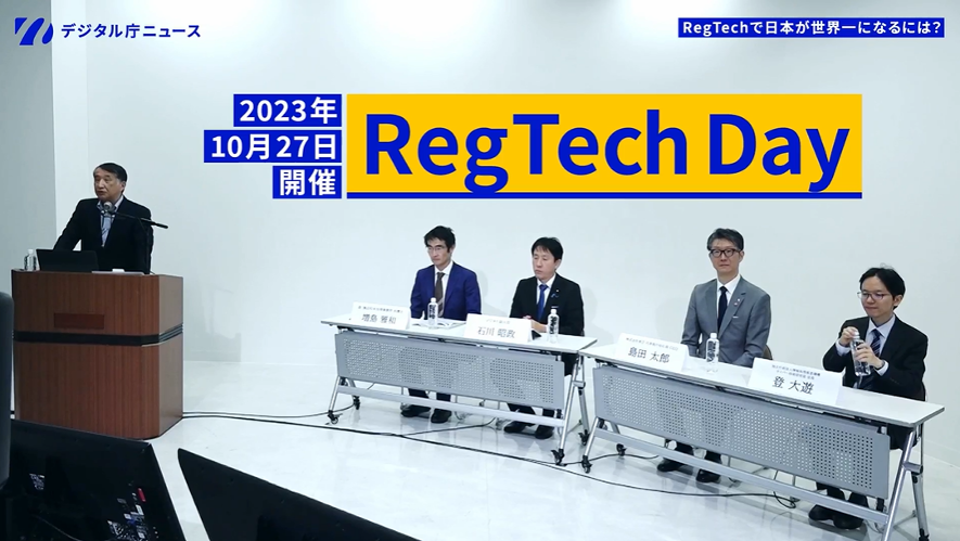 RegTech Dayの撮影会場の風景。左側に司会が演台を前に立っていて、右側に登壇者が4人テーブルを前にして座っている。登氏は登壇者の一番左側にいる。