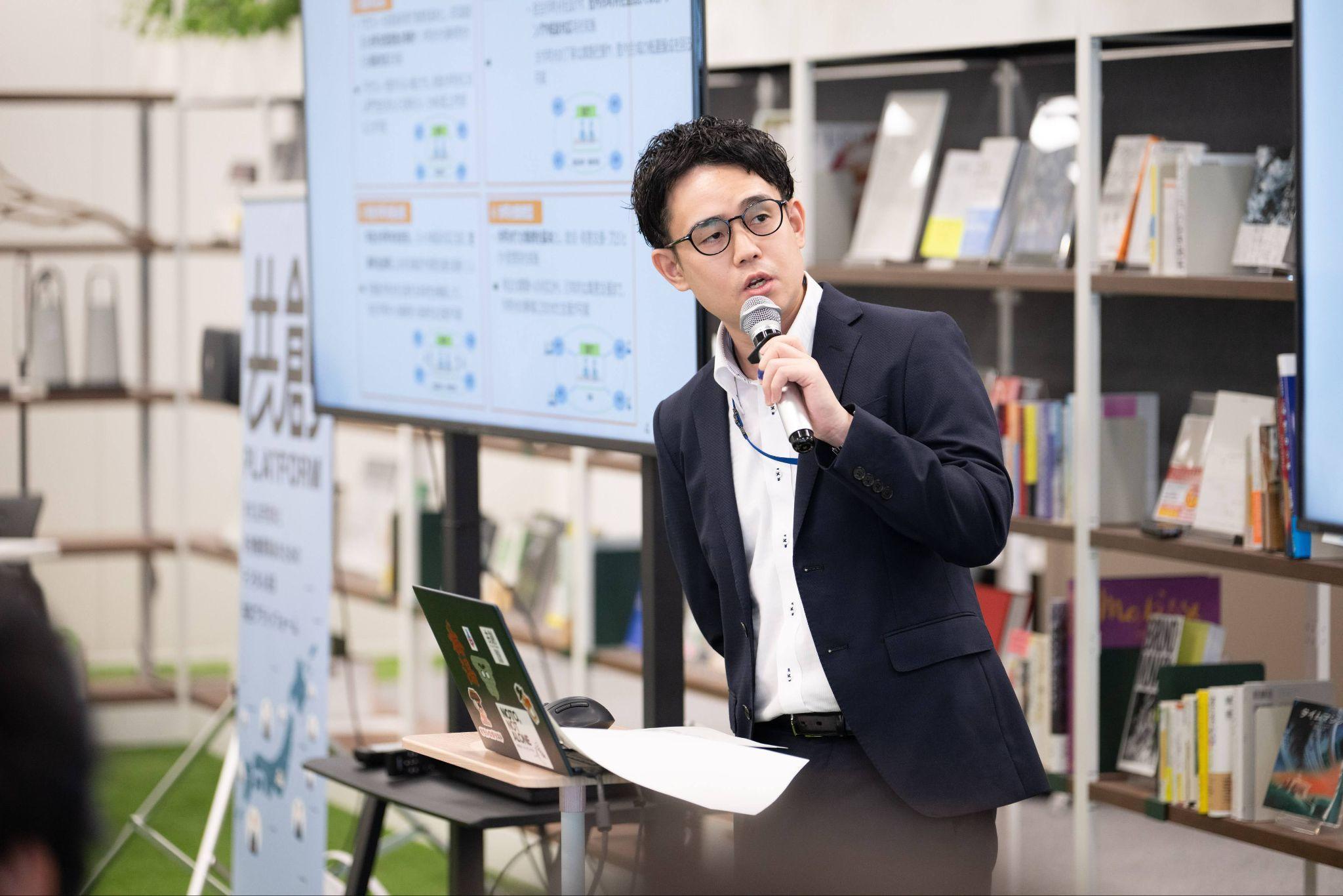 総務省の櫻井俊寿氏が左手にマイクを持ち話している様子。櫻井氏の前にはノートパソコン。背景にはモニターや、共創PFのパネル、そして会場のデジタル庁の芝生エリアにある本の数々がうっすらと見える。