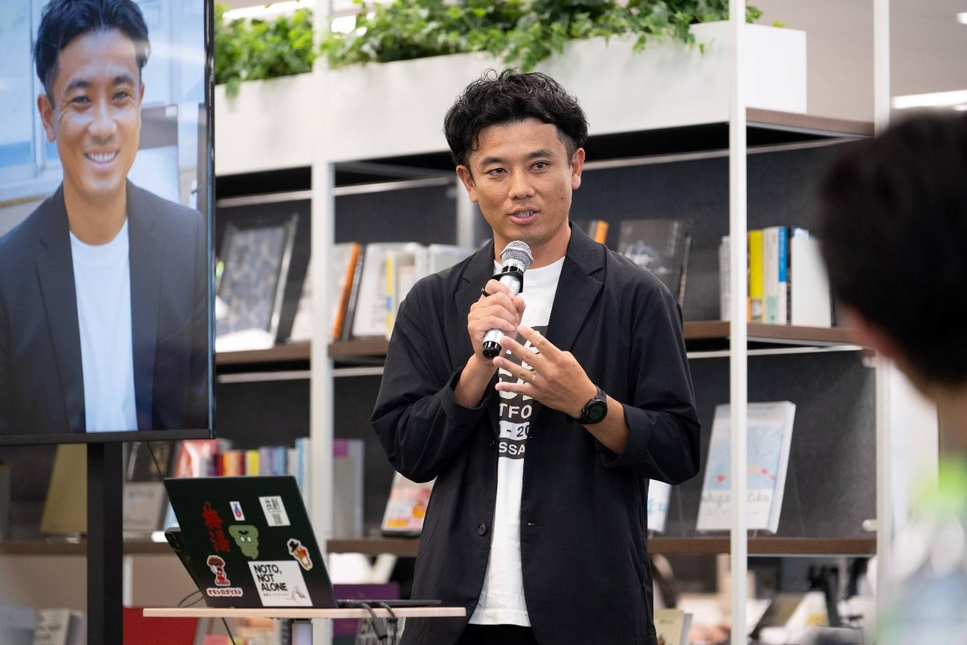 下呂市の長尾飛鳥氏が右手にマイクを持って話している様子。長尾氏の前にはノートパソコンがあり、後ろにはモニターが見切れている。モニターの画面には自身の自己紹介のスライドに掲載されていた顔写真が見える。