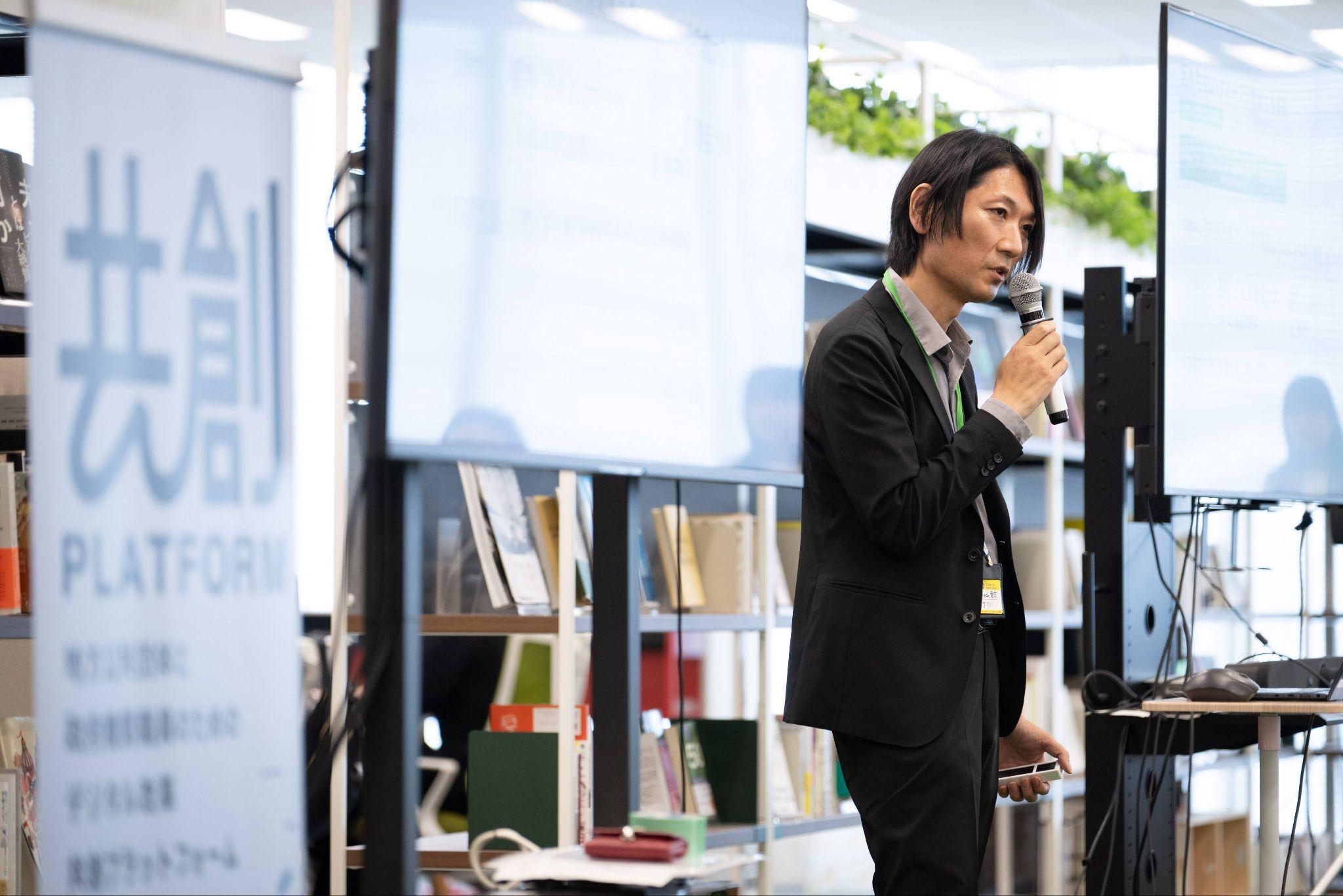 GovTech東京の田邊進一氏が右手にマイクを持って、立ちながら話している様子。田邊氏の両サイドにはモニターが置かれているが、文字は反射で見えない。写真の左側には「共創PLATFORM」と書かれたパネルも見えている。