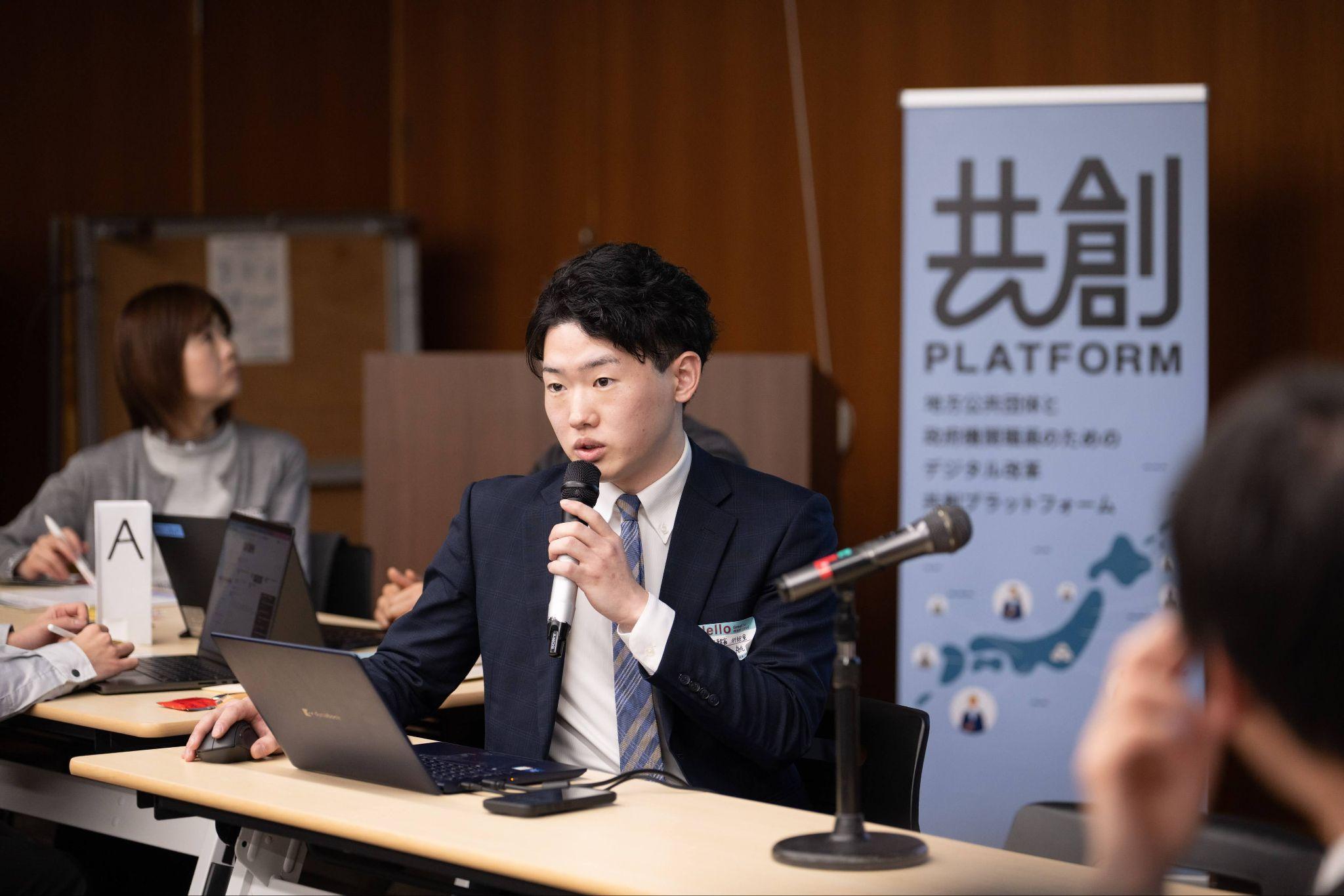 共創PLATFORMのイベントに登壇する総務省自治行政局市町村課行政経営支援室の岡野航大氏。テーブルの前に座りマイクを口元に近づけながら発言している。テーブルの上にはノートパソコンとスマートフォンが置かれている。右後方には『共創PLATFORM 地方公共団体と政府機関職員のためのデジタル改革 共創プラットフォーム』と書かれたロールアップバナーが立っている。