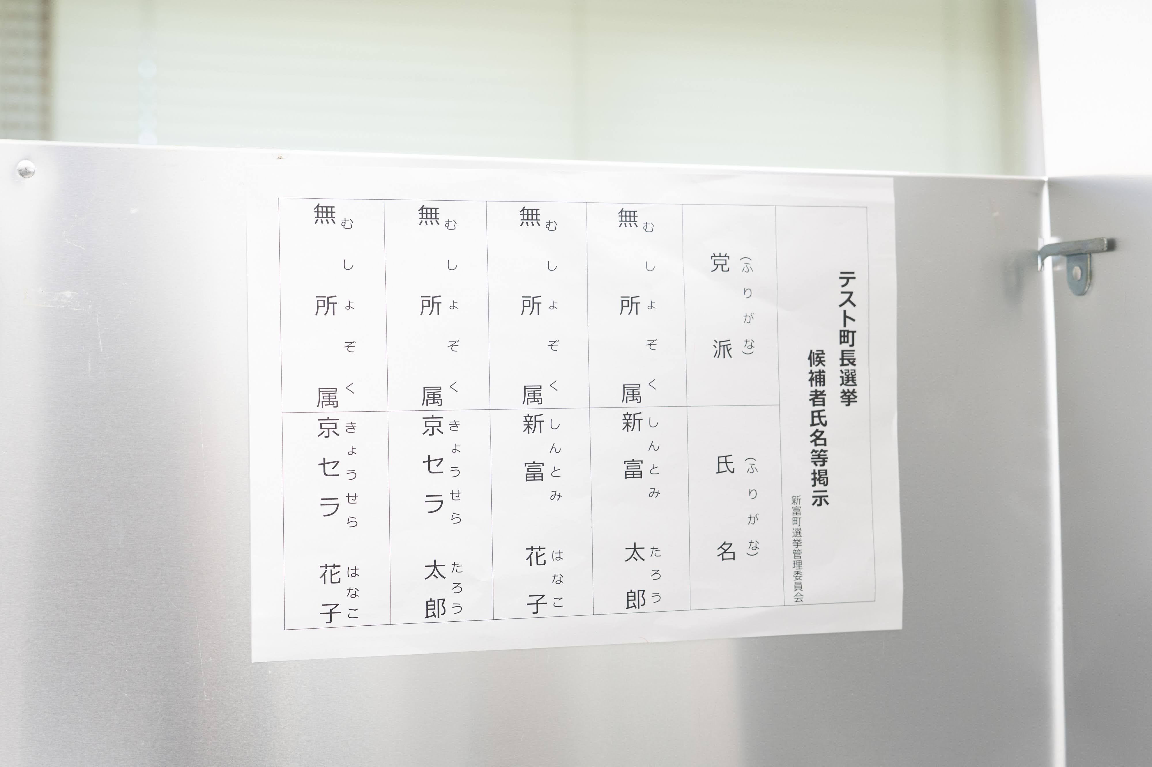 記載台に、候補者氏名等掲示の用紙が貼られている。テスト・実証実験用のダミーデータ。タイトルは「テスト町長選挙 候補者氏名等掲示 新富町選挙管理委員会」。表は5列構成で、一番右に「党名（ふりがな）」「氏名（ふりがな）」と書かれている。左から「無所属 京セラ 花子」「無所属 京セラ 太郎」、「無所属 新富 花子」「無所属 新富 太郎」と記載されている。